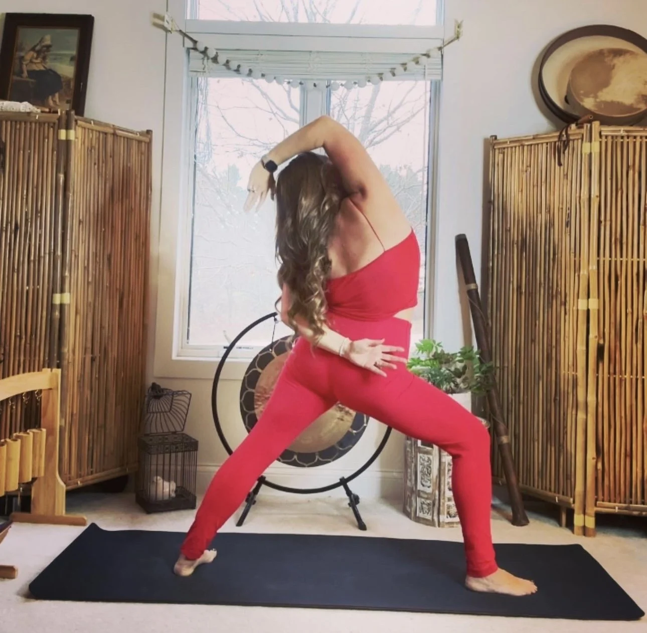 Woman practicing yoga in a living room with bamboo room dividers, a gong, and a window with trees outside.