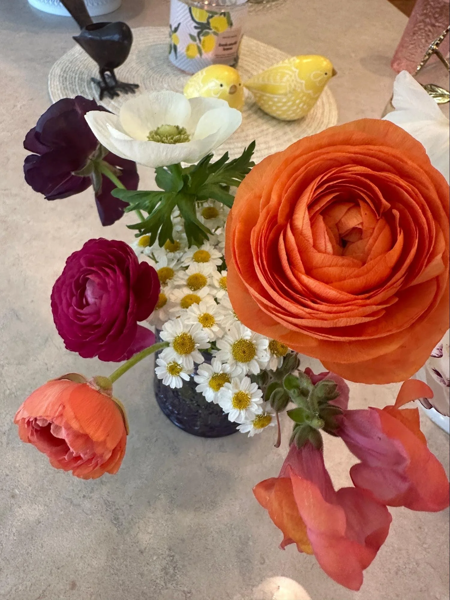 Close-up of a floral arrangement with orange ranunculus, white anemone, purple and pink ranunculus, small white daisies, and yellow billy buttons in a dark vase, with decorative yellow bird figurines and a decorative can with lemon print in the backg