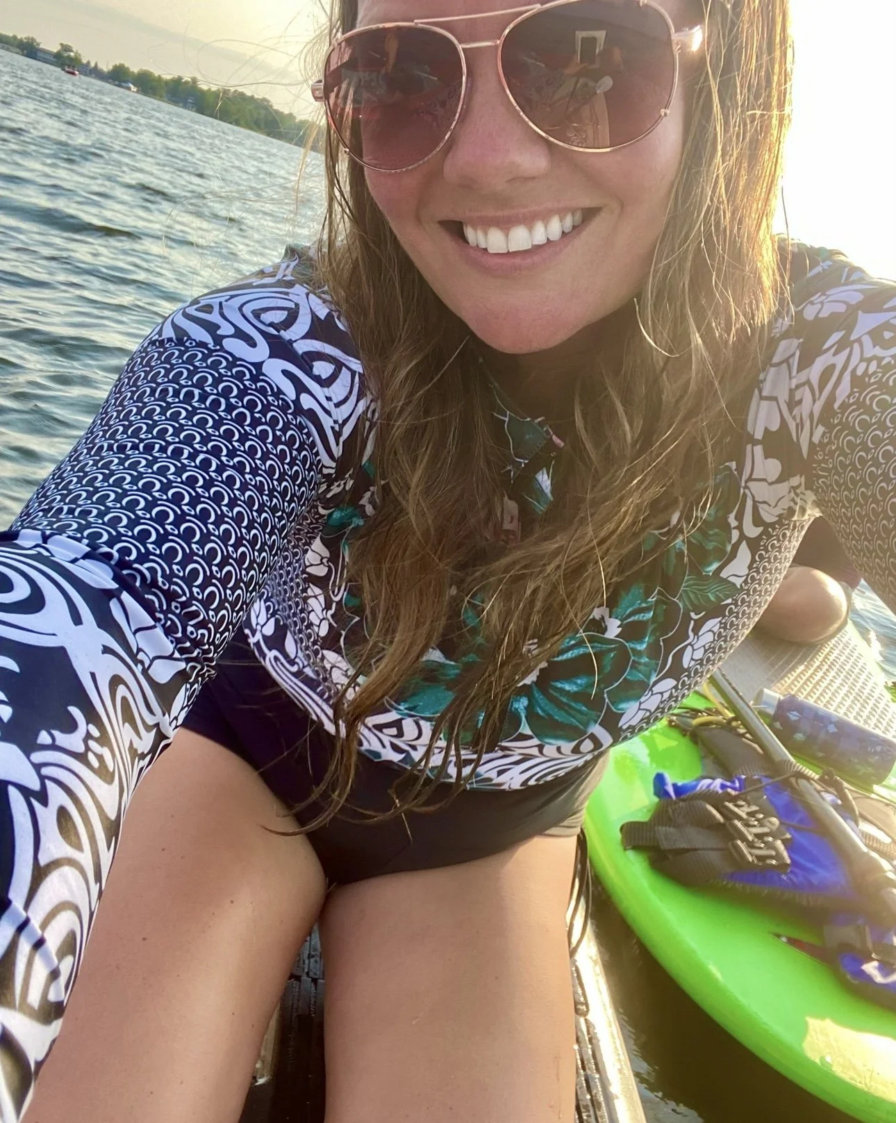 A woman smiling on a paddleboard on a lake, wearing sunglasses and a patterned long-sleeve shirt, with water and trees in the background.