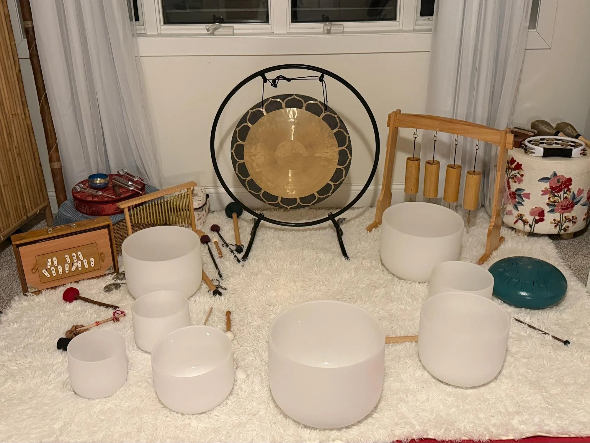 A room with a large gong, wind chimes, and various musical instruments on a fluffy white rug. There are white curtains, a window with a white frame, and a variety of objects including drums, tambourines, and a colorful decorative pillow.