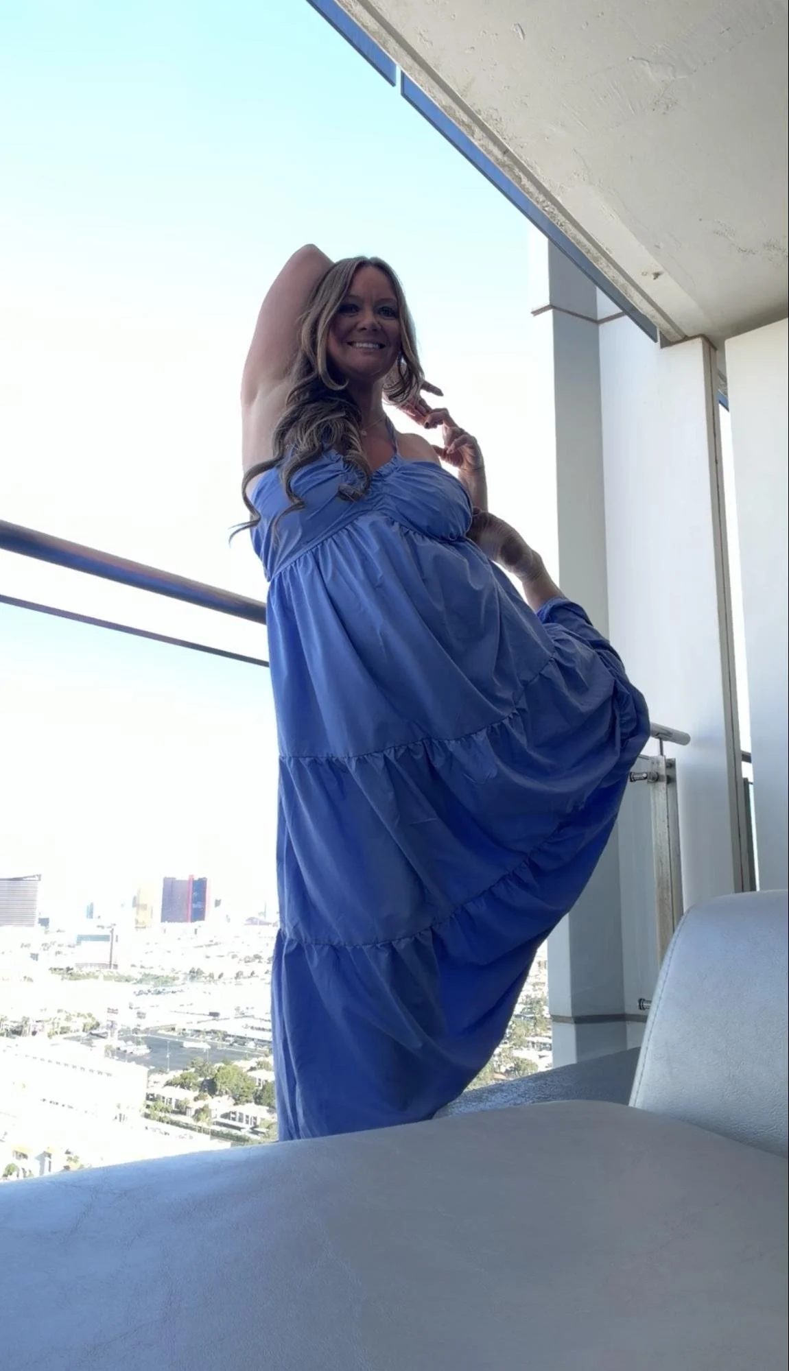 Woman in a blue dress standing on a balcony with a city view and clear sky.
