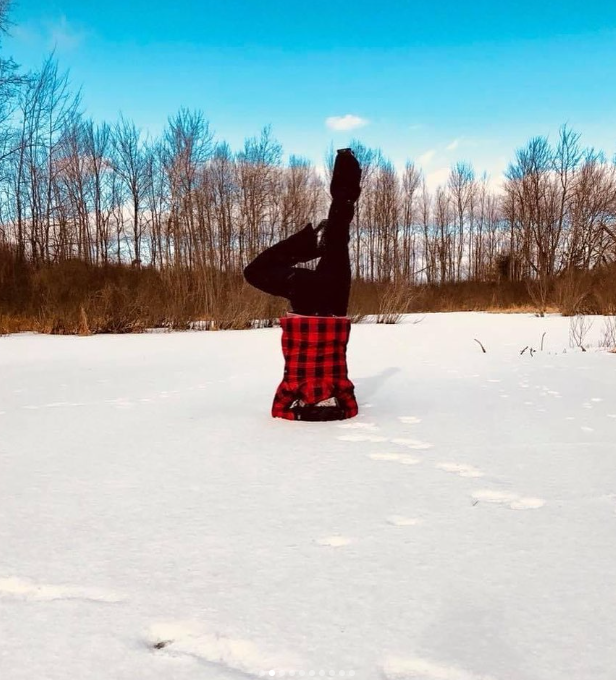 Person doing a headstand in a snowy landscape, wearing a red and black checkered jacket and black pants, with footprints leading to their position, and trees and a blue sky in the background.
