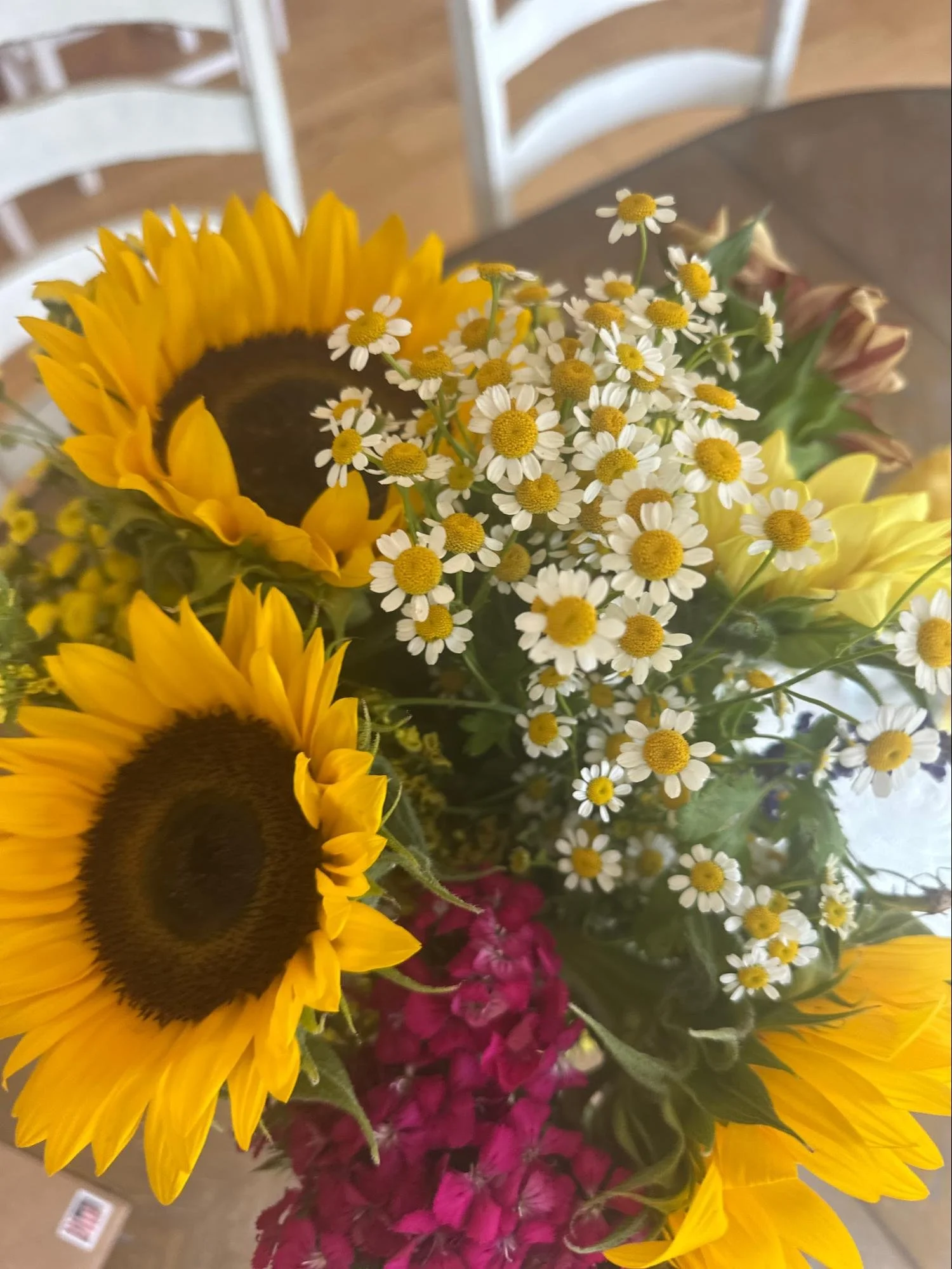 Arrangement of sunflowers, daisies, pink flowers, and greenery on a table with white chairs in the background.