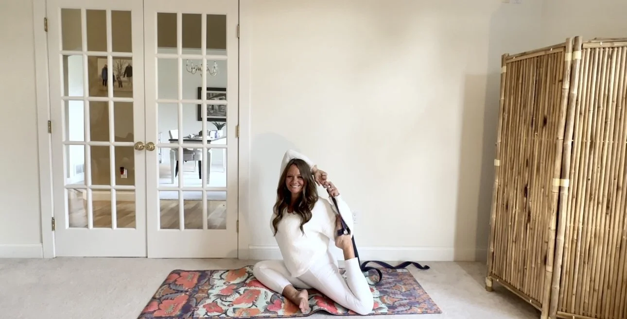 A woman with long brown hair smiling and sitting on a colorful rug, wearing a white hoodie and white leggings, in a room with glass double doors and a bamboo storage unit.