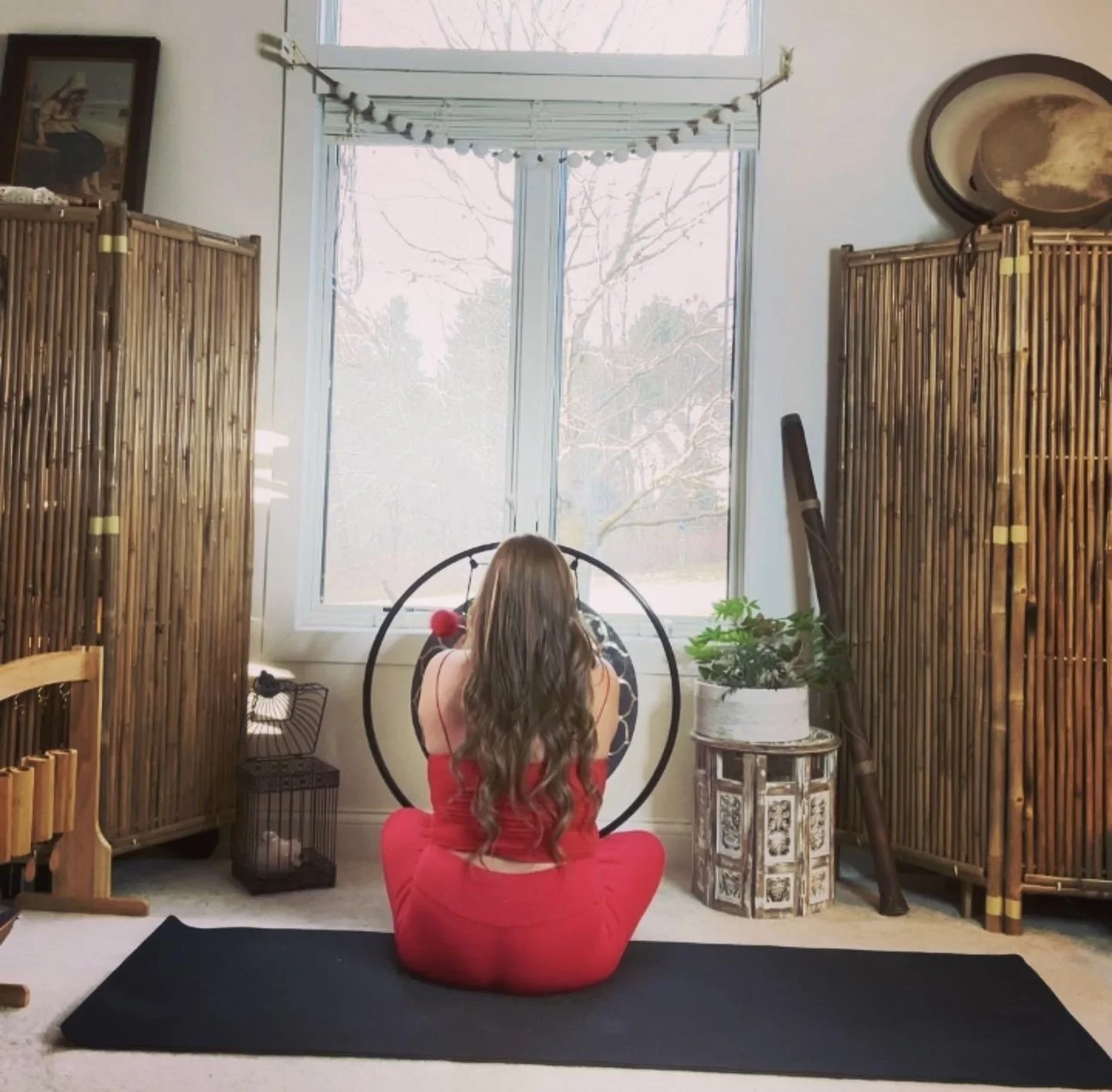 A woman in a red dress practicing yoga in a bright room with large window, surrounded by wooden screens, houseplants, and decorative items.