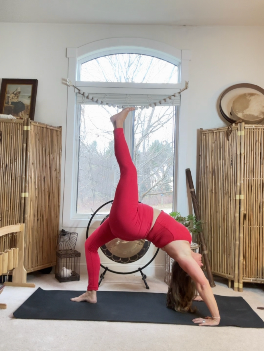 A woman in red leggings and top performing a yoga pose called downward dog in a room with bamboo furniture and a large window behind her.