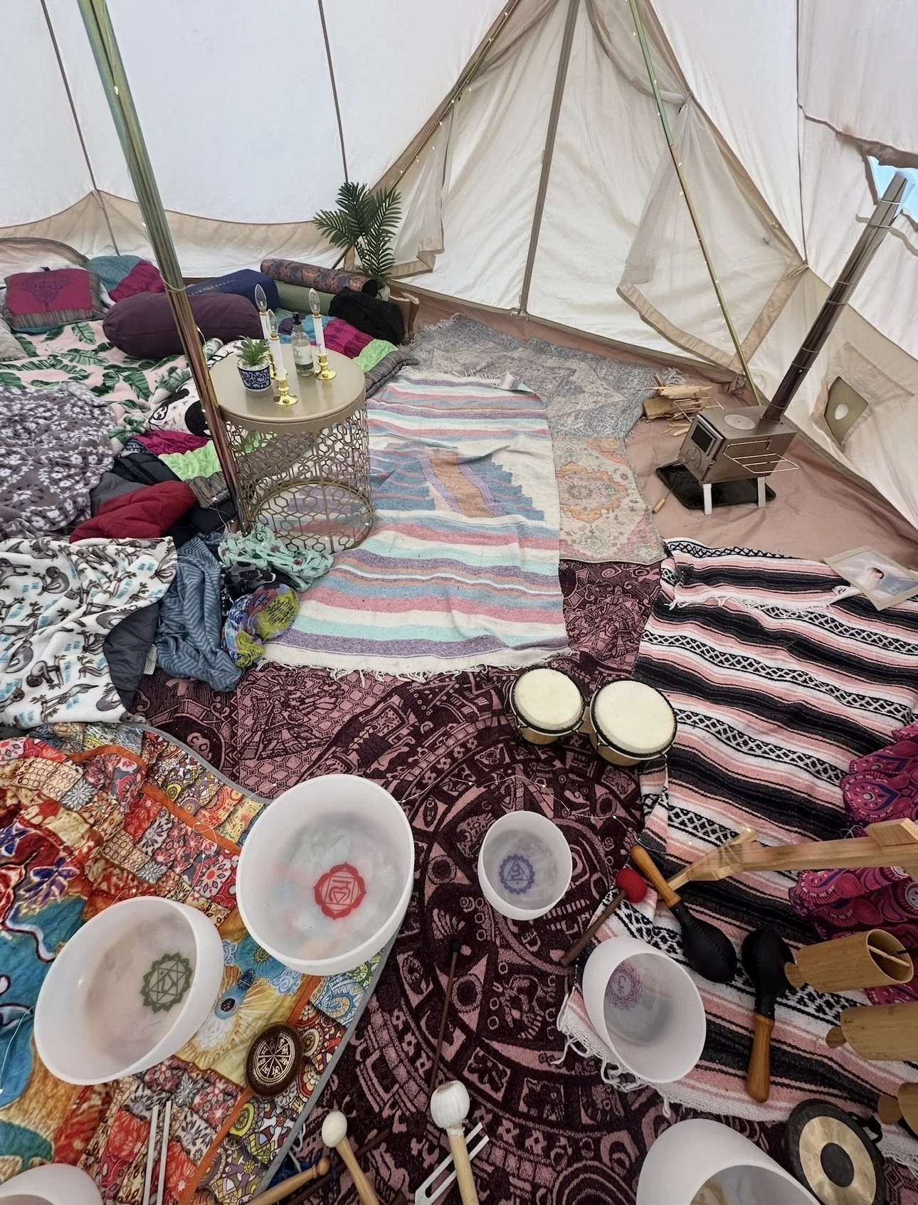 Inside a cozy tent decorated with colorful rugs, towels, and cushions, featuring a small table with plants and candles, a heater, and percussion instruments like drums and bowls.