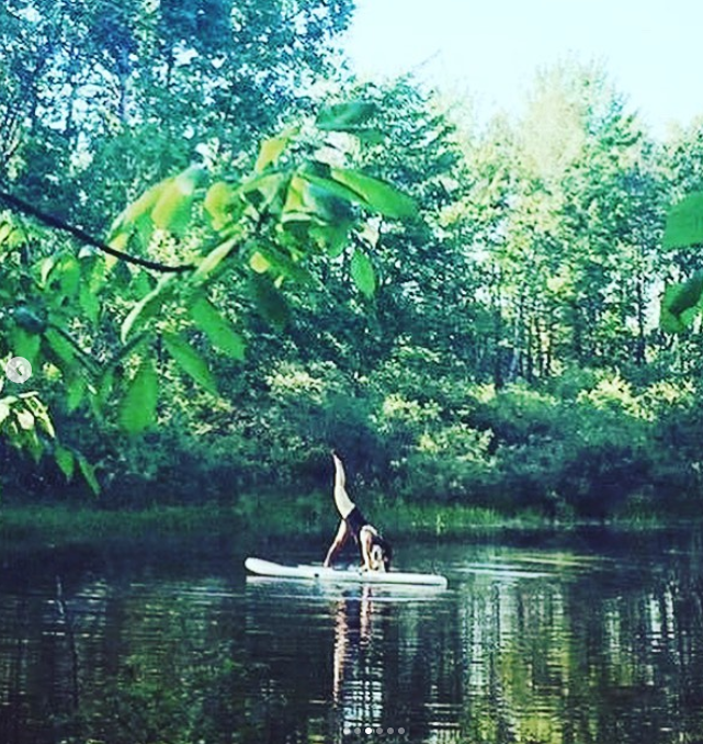 Person doing a handstand on a paddleboard on a calm river in a lush green forest.