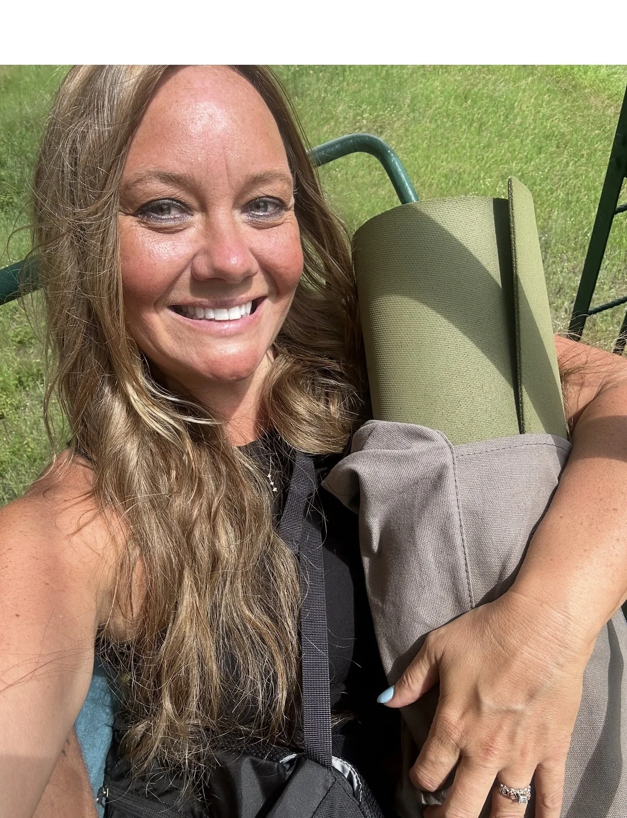 Woman with long curly hair smiling outdoors, holding a large bag with a rolled-up yoga mat, sitting on a swing on a grassy area.