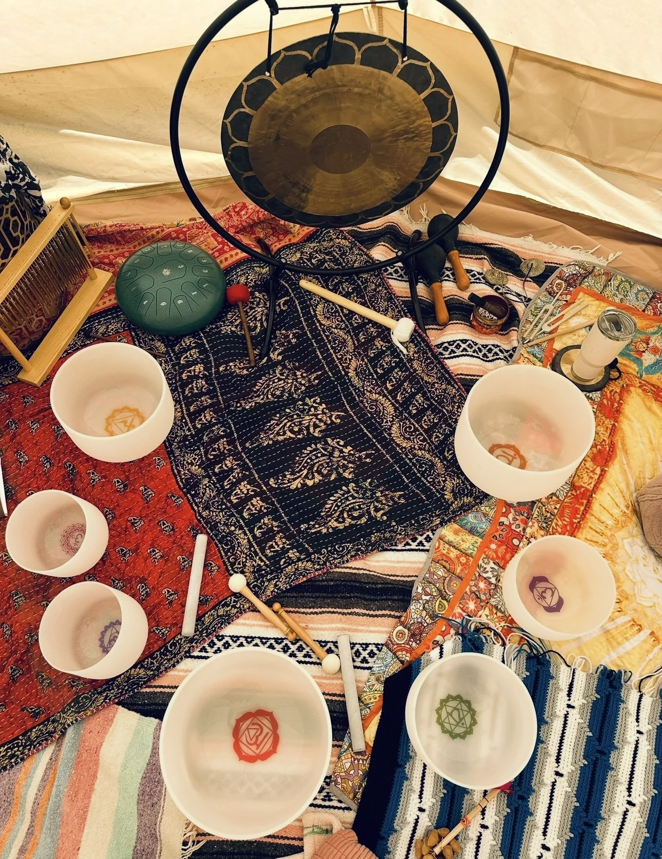 Arrangement of Tibetan singing bowls, gongs, mallets, and a percussion instrument on colorful fabric with a backdrop of a tent interior.