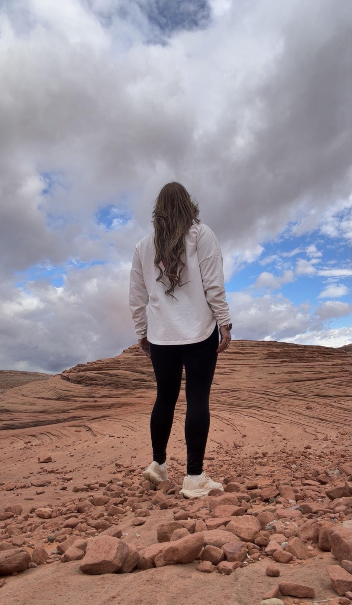 A woman with long brown hair in a white jacket, black leggings, and white sneakers standing on rocky terrain under a partly cloudy sky.