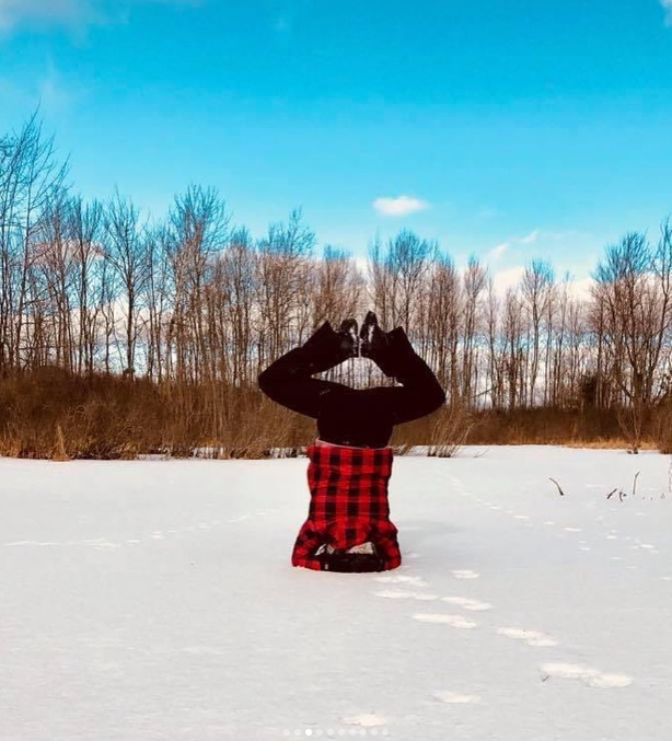 Person in a red and black plaid shirt doing a headstand on snow with bare trees and a blue sky in the background.