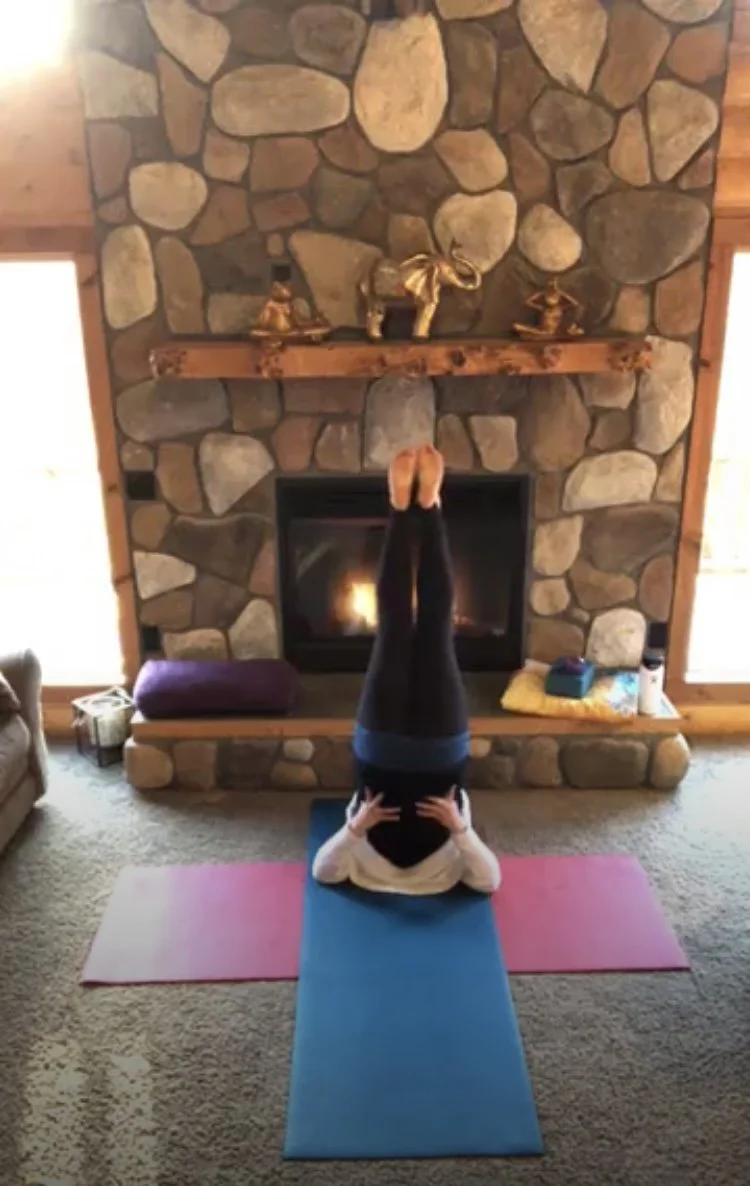 Person practicing yoga or meditation in a living room, lying on a green yoga mat with head resting on hands, in front of a stone fireplace with decorative brass animal sculptures on the mantle.