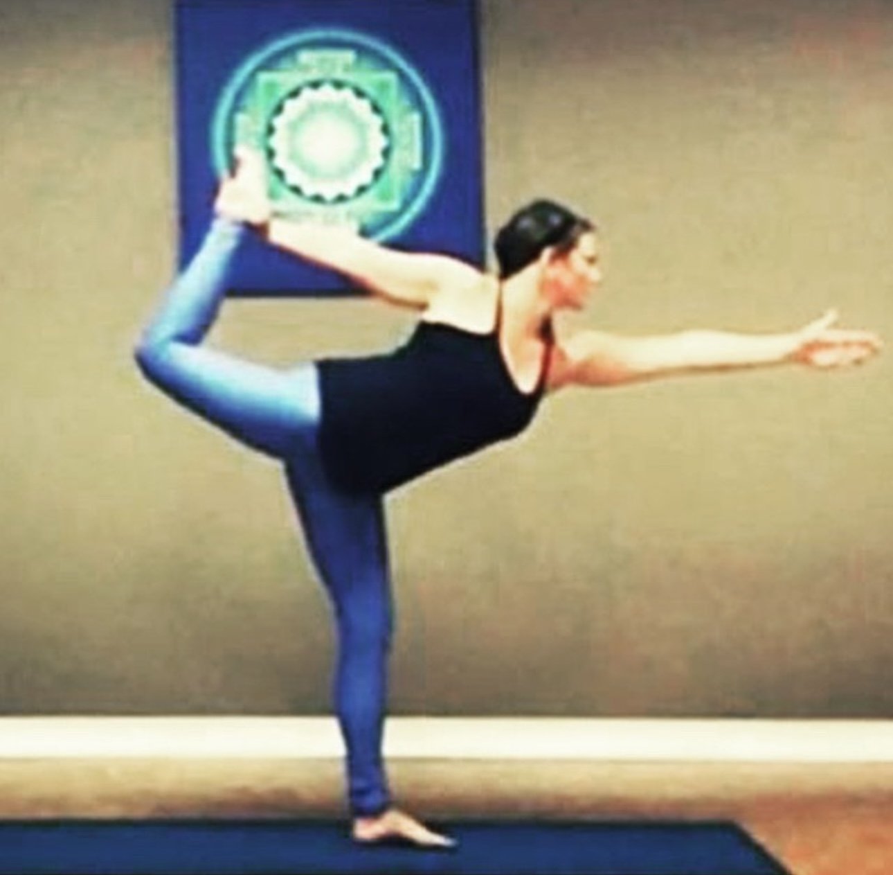 A woman practicing yoga indoors, standing on one leg while holding the other leg behind her with one hand and extending her other arm forward.