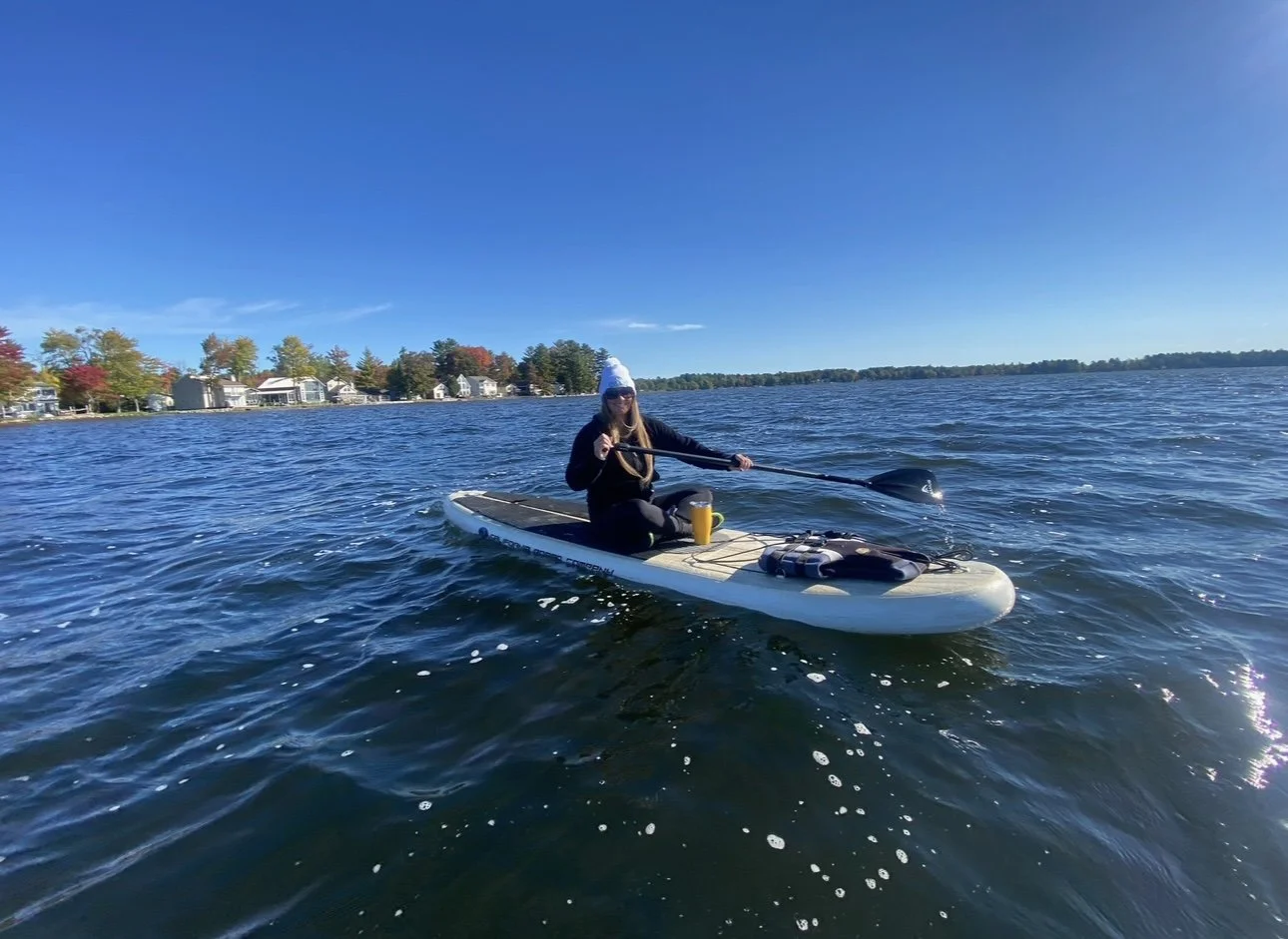 A person wearing sunglasses and a white beanie sitting on a paddleboard on a lake, holding a paddle, with houses and trees in the background under a clear blue sky.