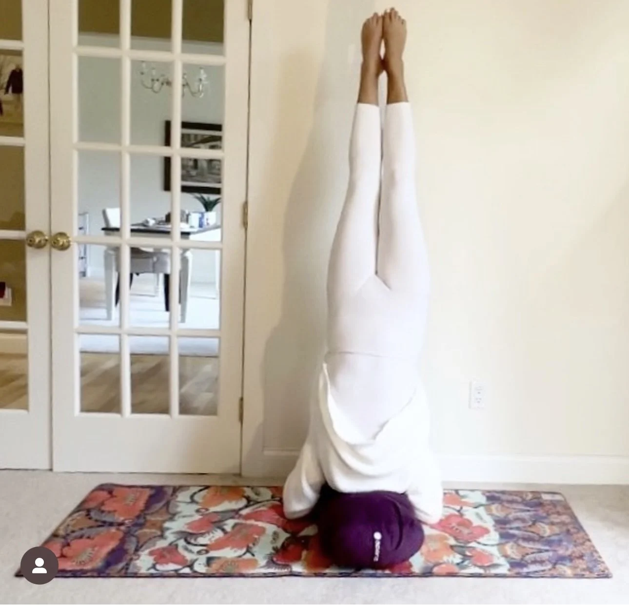 A person practicing a headstand yoga pose on a colorful floral rug in a room with a door and a dining area visible through the glass panes.