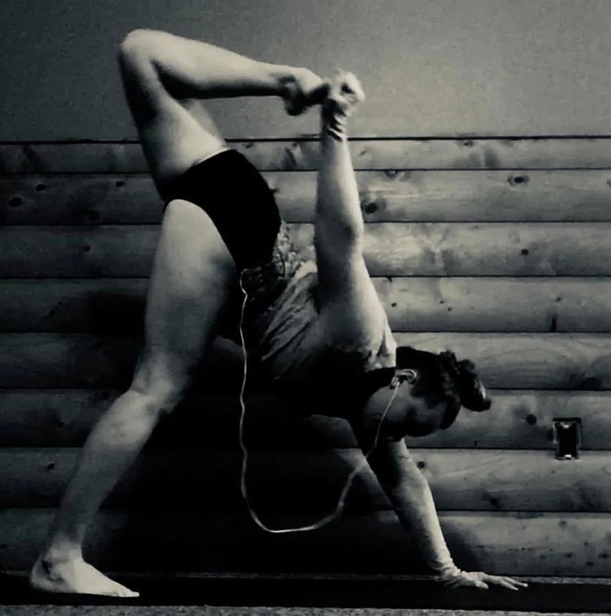 Person practicing yoga in a handstand pose against a wooden wall, wearing athletic clothing and earphones.