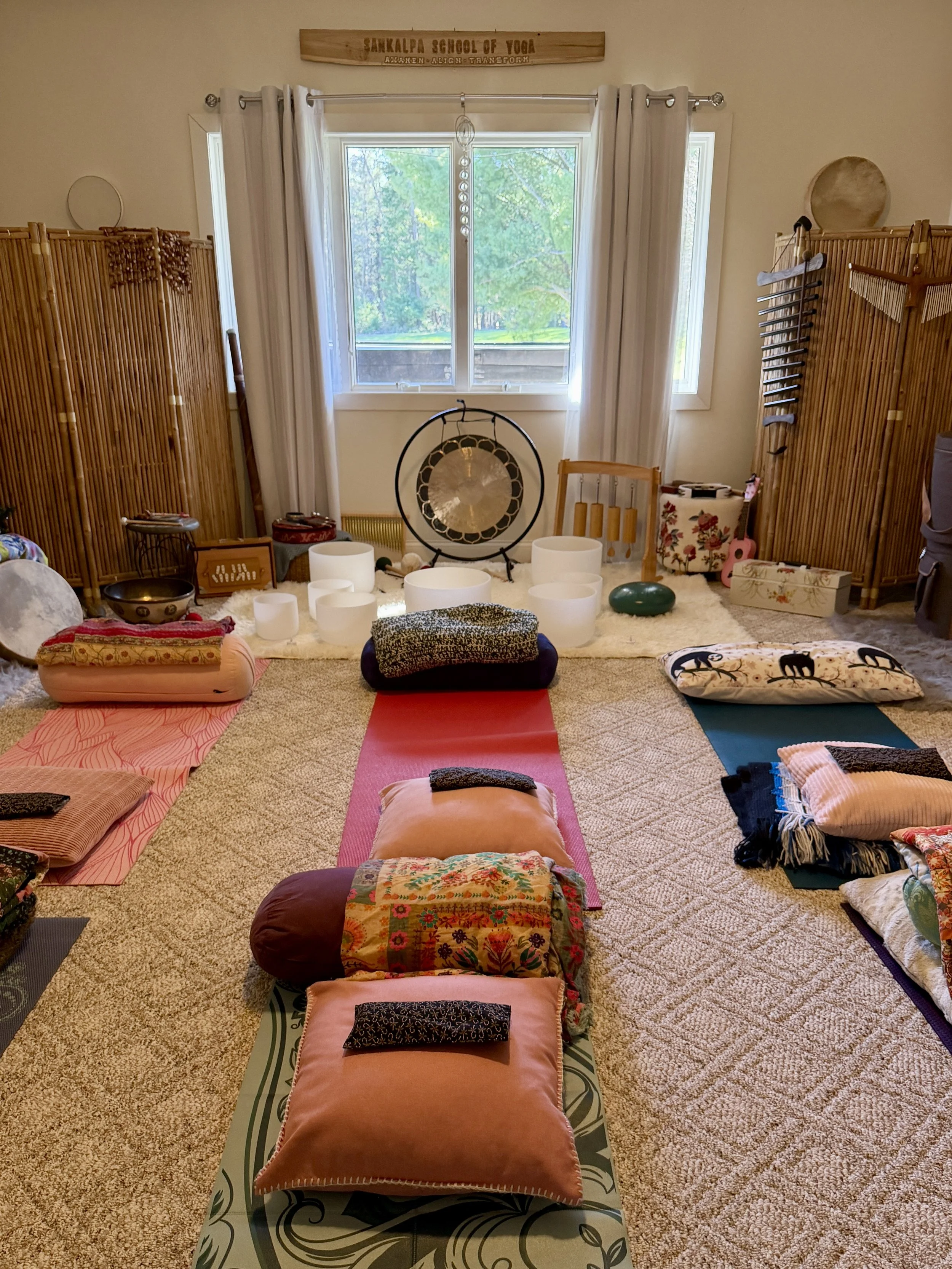 A cozy indoor space set up for a yoga or meditation session with mats, cushions, and a gong, near large open windows revealing a green outdoor view, with wooden room dividers and spiritual decor.