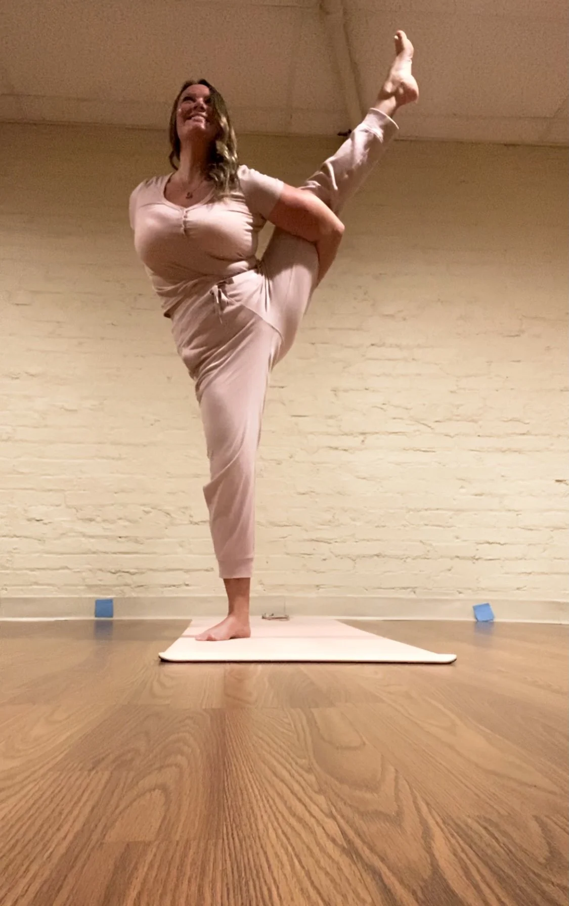 Woman practicing yoga on a mat in a room with wooden floors and a beige brick wall. She is balancing on one foot while holding her other leg behind her in a stretch, smiling.