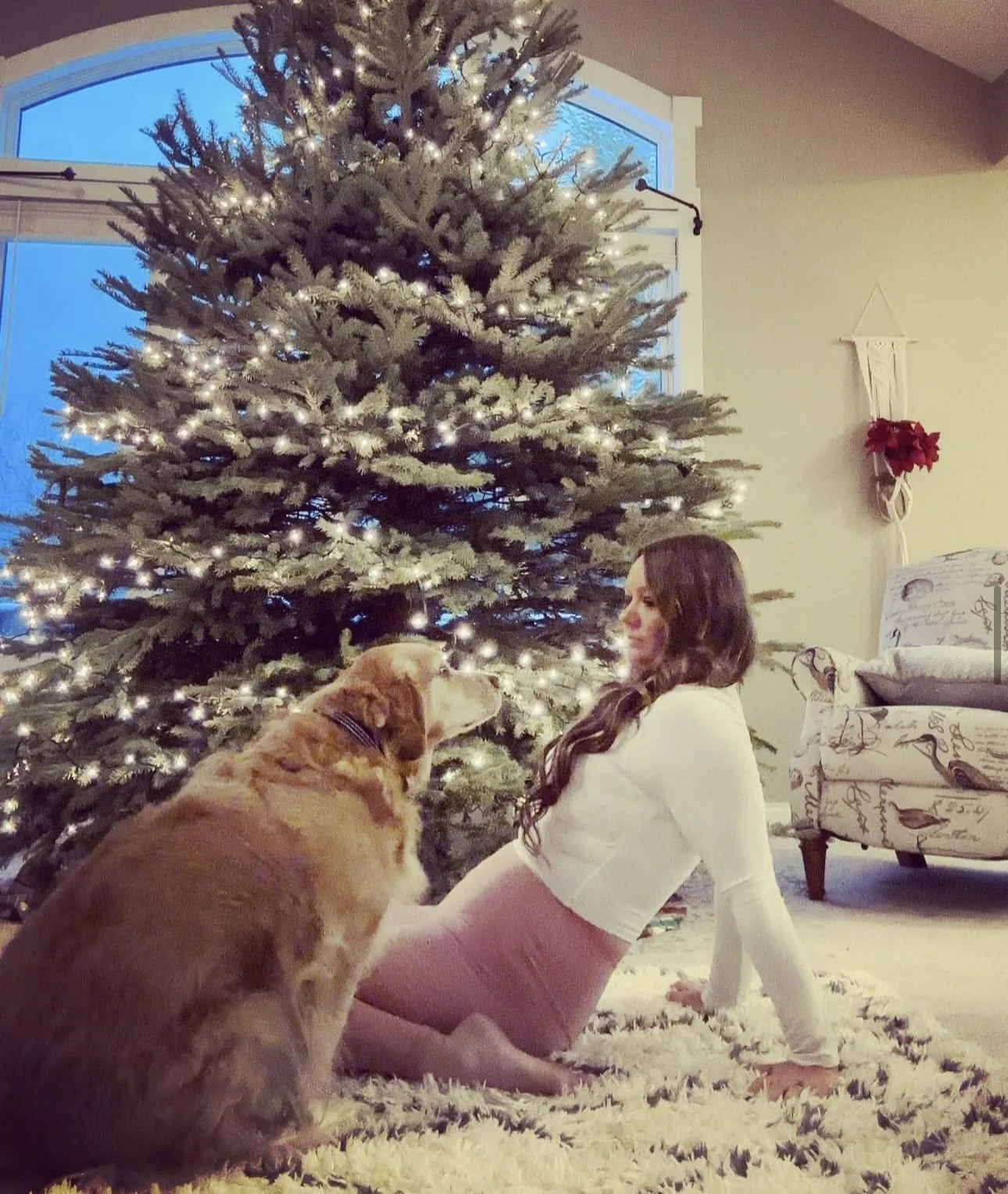 A woman with long hair, wearing a white top and pink pants, is sitting on a fluffy rug in front of a decorated Christmas tree with white lights. She is interacting with a large dog, likely a retriever, that is sitting beside her, and they are touchin