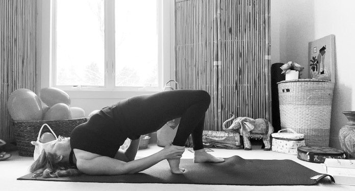 A woman practicing yoga in a room, in a bridge pose on a mat, with large windows and decorative items in the background.