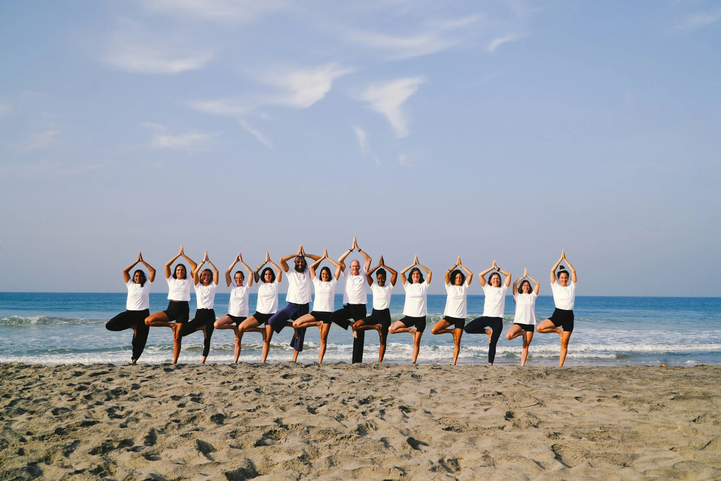 A group of people, including men and women, practicing yoga on the beach during daytime. They are standing in a line with their hands joined above their heads in a yoga pose, against the backdrop of the ocean and sky.