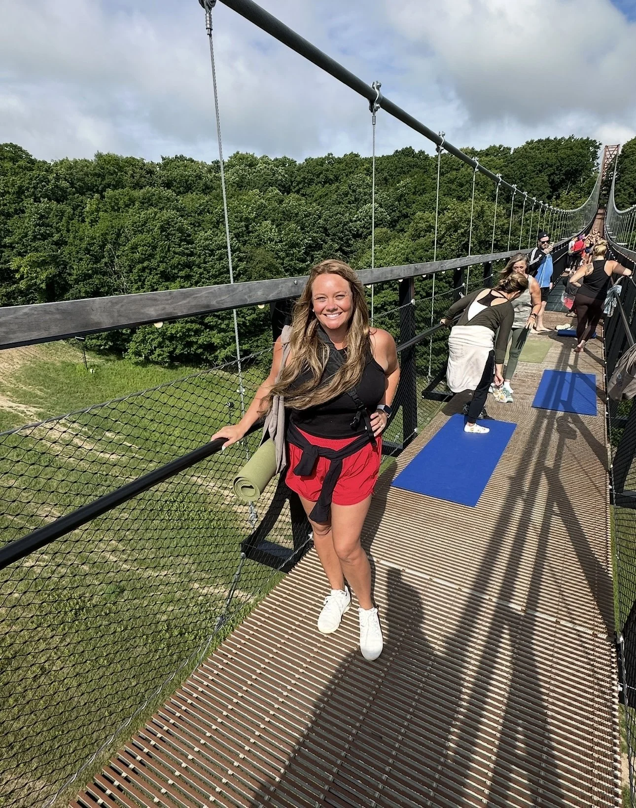 A woman standing on a suspension bridge with a forest green landscape in the background, participating in a group yoga session with others on yoga mats.