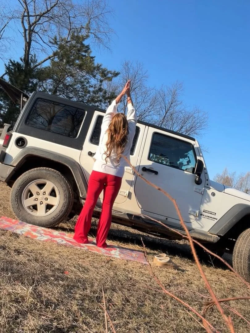 A woman with long, wavy brown hair is standing on a red and white patterned blanket on the ground, next to a white Jeep. She is wearing red pants and a white top, and has her arms raised above her head, palms pressed together. The woman is outdoors o