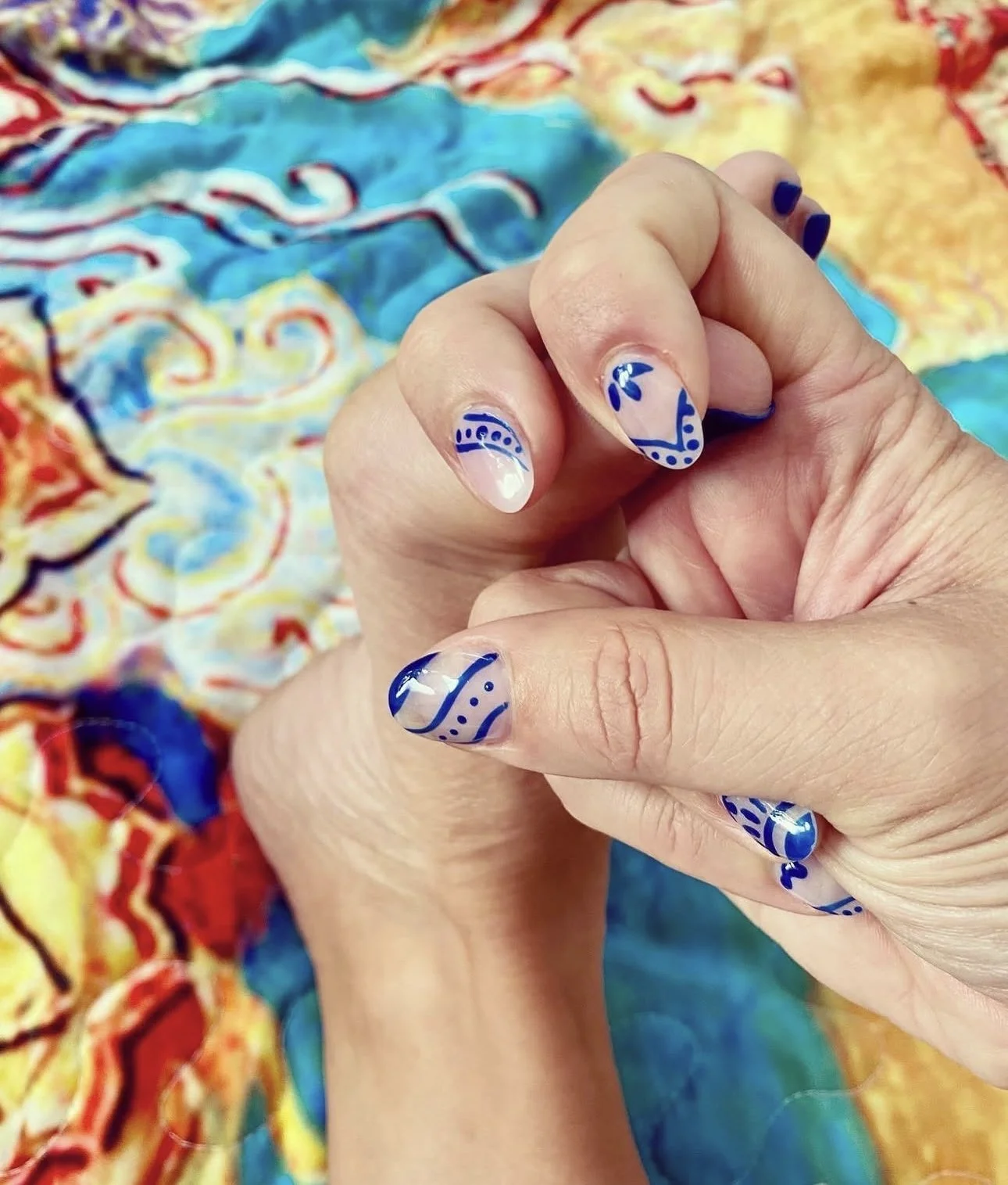 Close-up of a hand with manicured nails painted in navy blue and white, featuring intricate abstract patterns. The background is a colorful, swirling fabric in shades of blue, yellow, red, and orange.