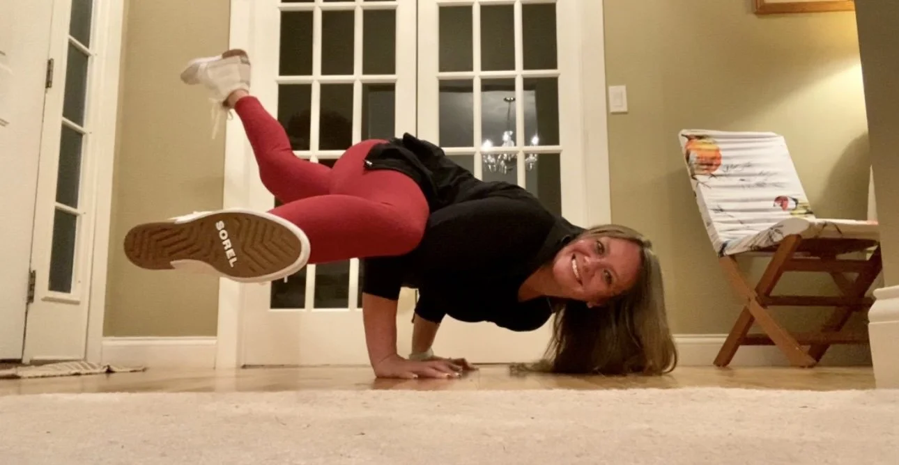 A woman wearing a black shirt and red leggings performing a handstand in a room with a glass door and a chair in the background.