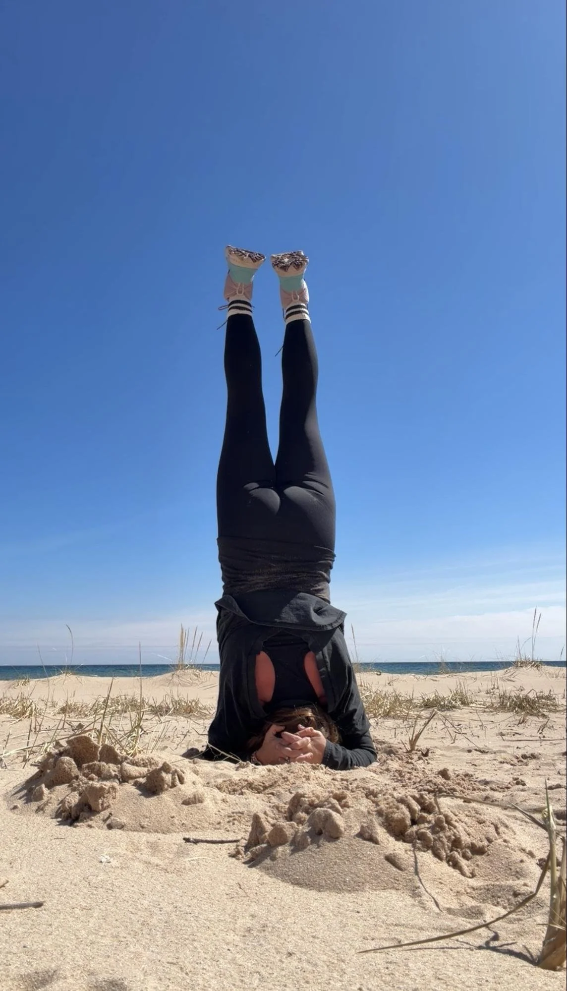 A person performing a headstand on a sandy beach, with their legs extended upwards and wearing black leggings, sneakers, and a dark jacket, under a clear blue sky.