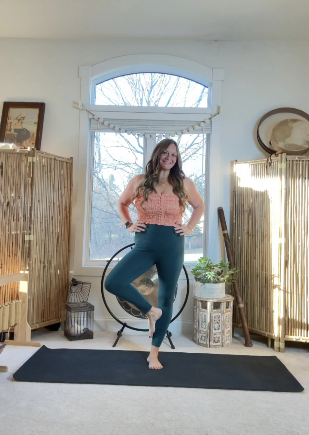 Woman practicing yoga in a sunlit room, standing on one foot within a circle apparatus, smiling, with decorative plants, bamboo screens, and artwork in the background.