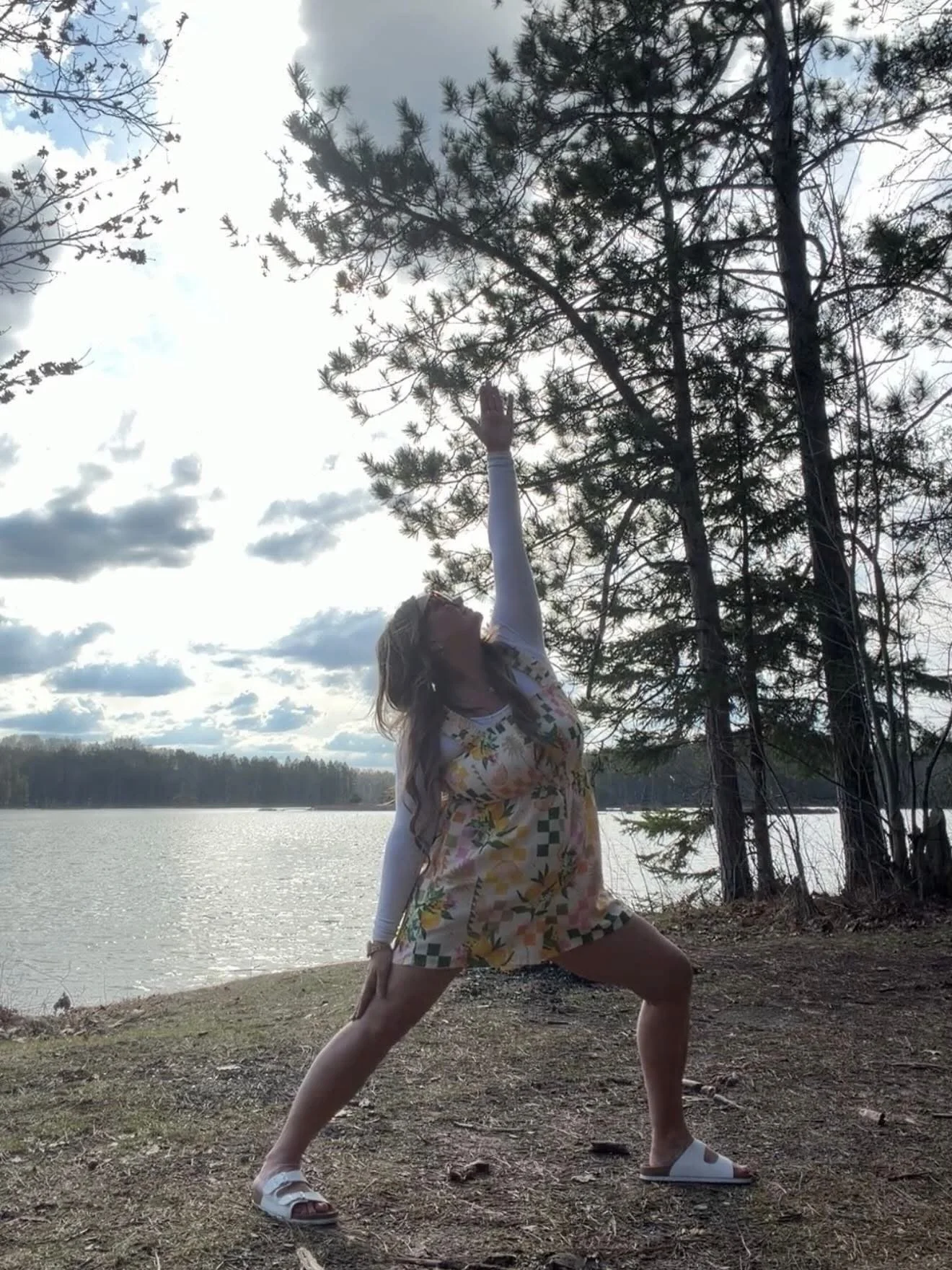 A woman in a yoga pose outdoors near a lake, with trees and cloudy sky in the background.