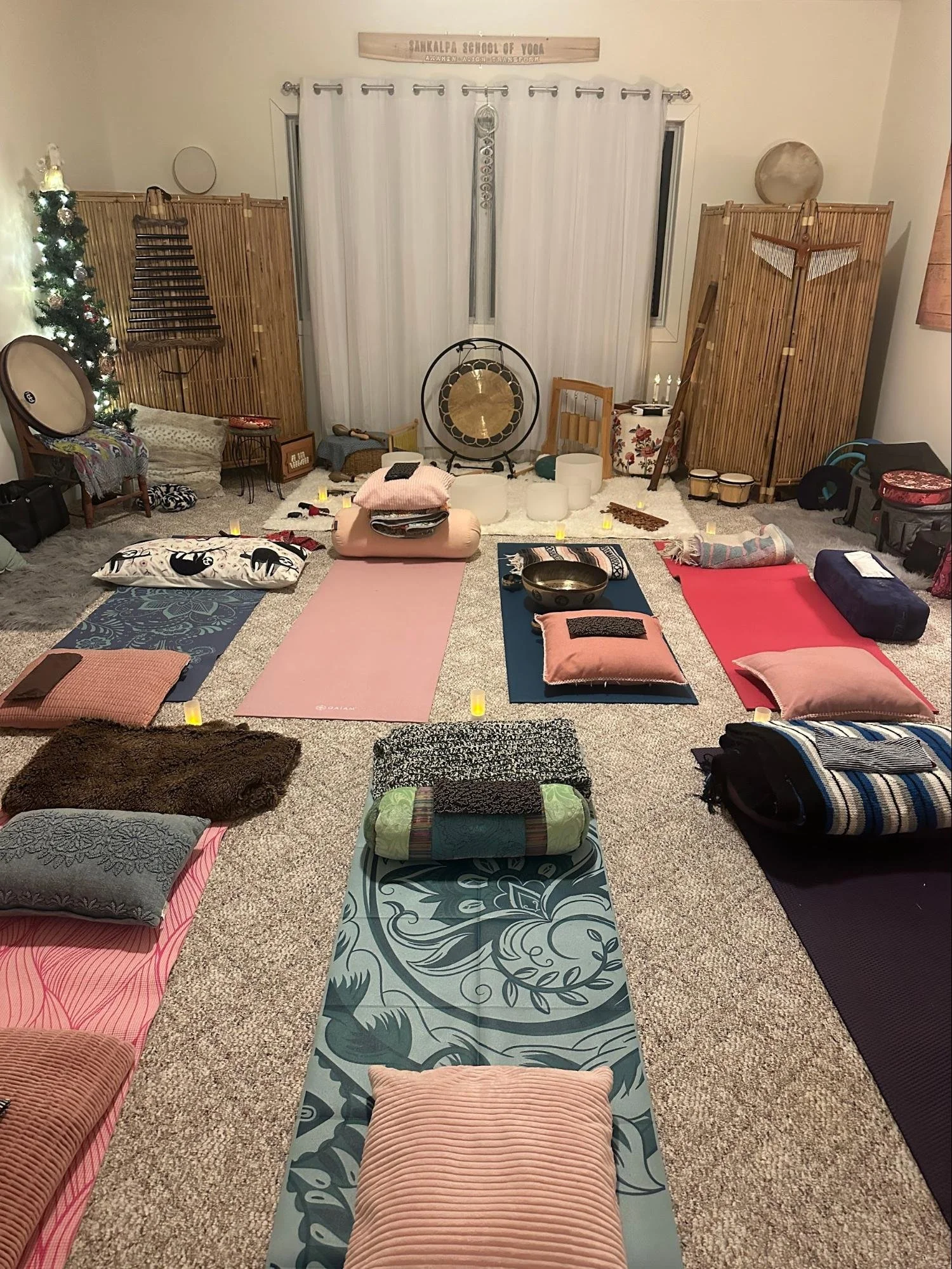 Room set up for a sound healing or meditation session with yoga mats, pillows, blankets, and candles on carpeted floor. There are bamboo room dividers, a Christmas tree with lights, and a gong in the background.