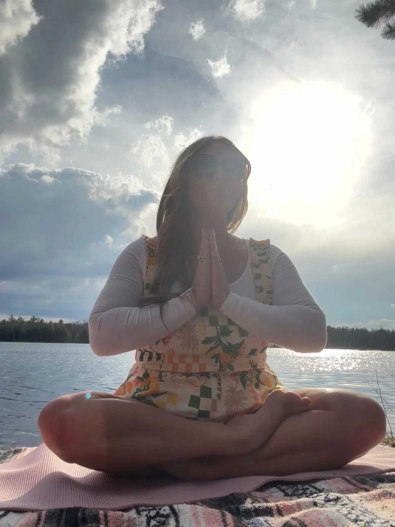 A woman practicing yoga outdoors by a lake, sitting cross-legged on a mat with her hands in prayer position, backlit by the sun and cloudy sky.