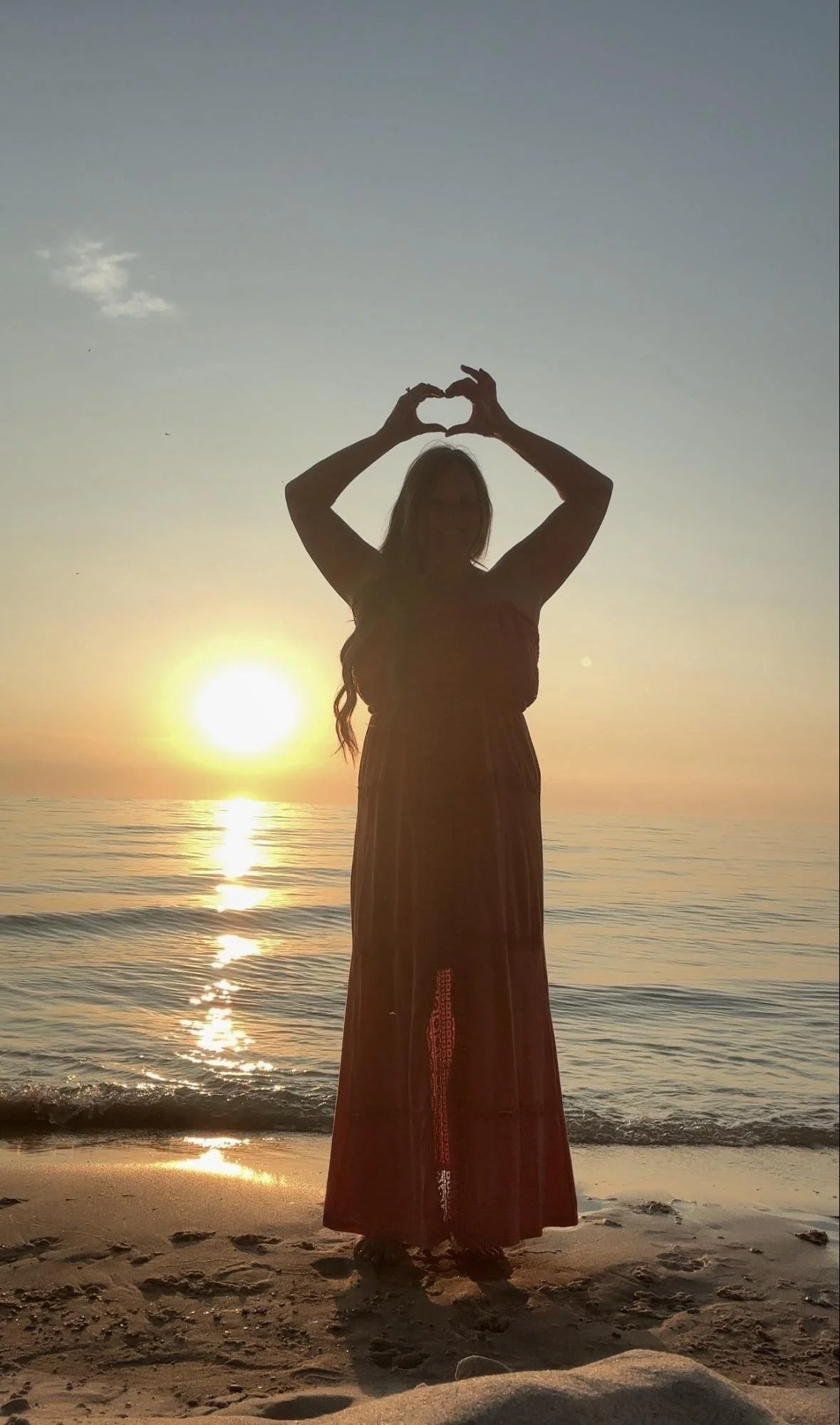 A woman standing on the beach at sunset, making a heart shape with her hands above her head. The sun is setting over the ocean behind her.