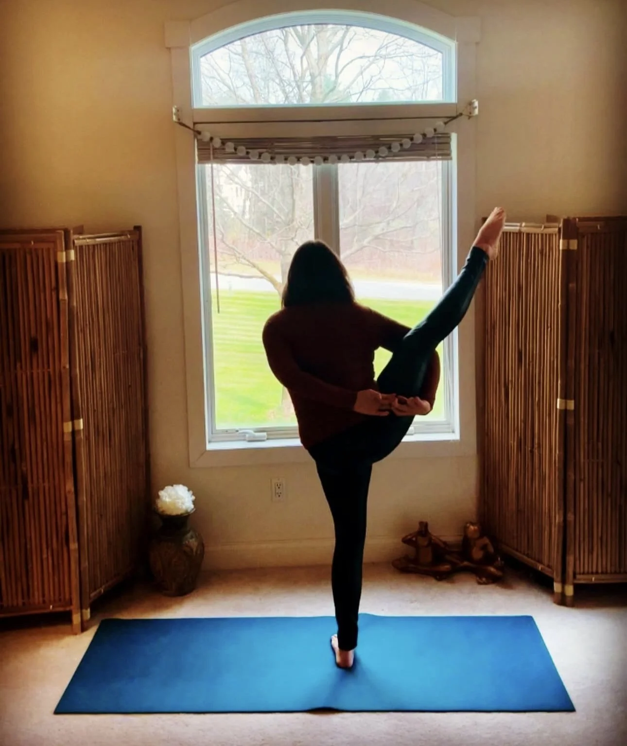 Person practicing a yoga pose on a blue mat indoors near a large window with curtains, wooden room dividers on sides, a vase with white flowers, displayed in a natural setting with trees outside.