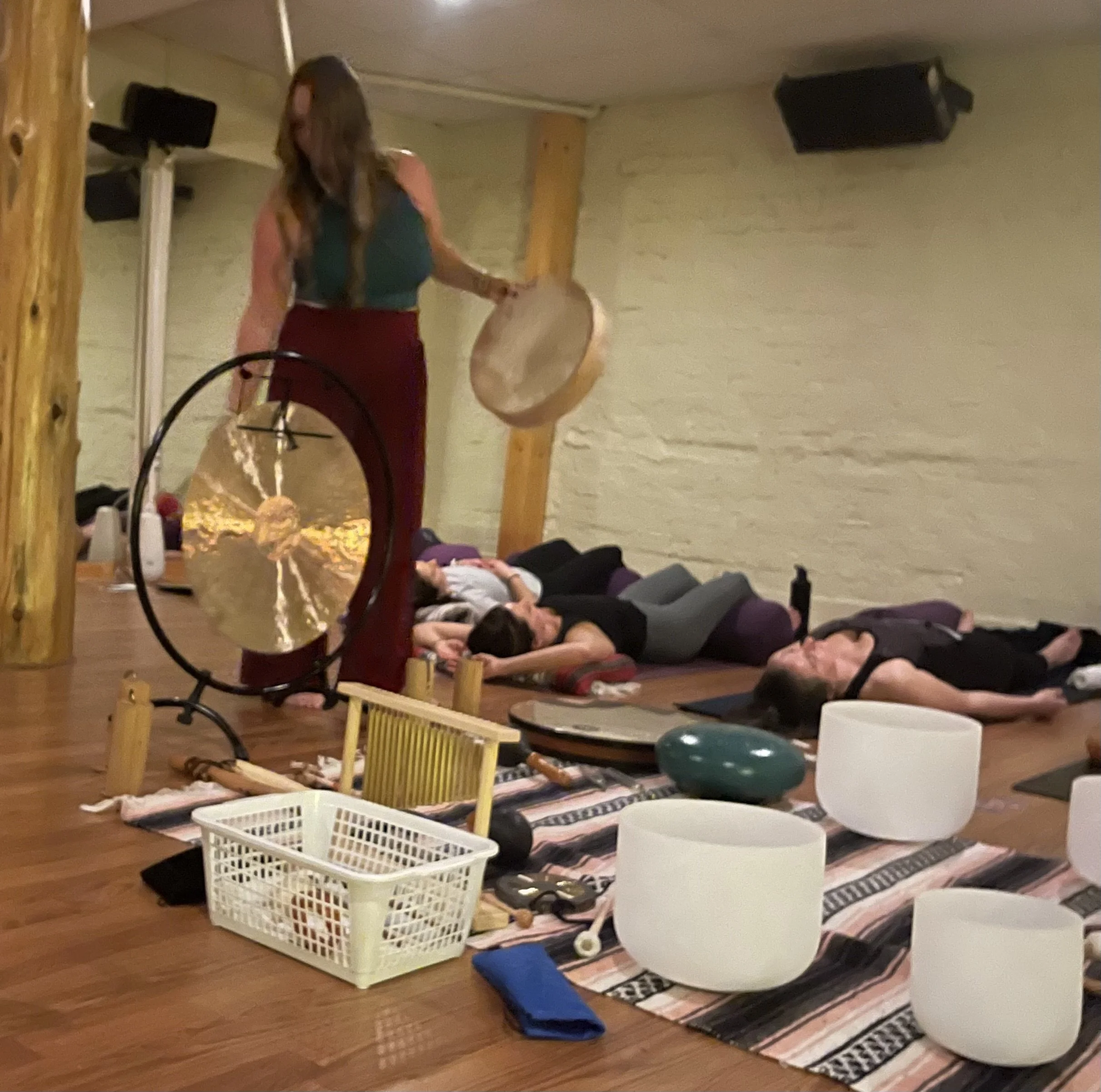 People lying on yoga mats during a class in a cozy room with beige brick walls. An instructor stands at the front near a drum, with speakers mounted on the wall.