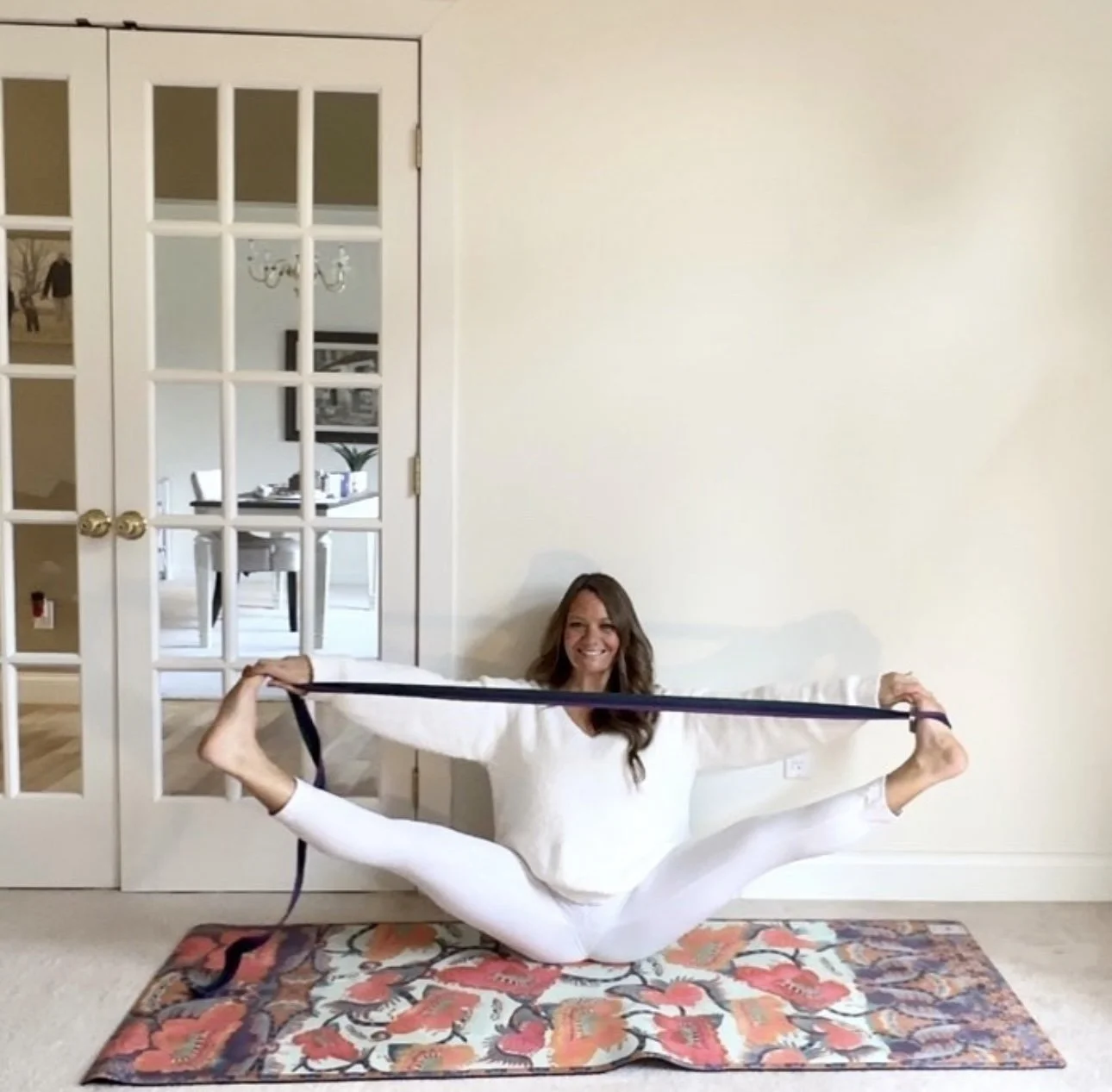 Woman in white Yoga clothes performing an extended side split on a colorful floral yoga mat, holding a yoga strap in a room with a glass-paneled door and a glimpse of a dining area in the background.