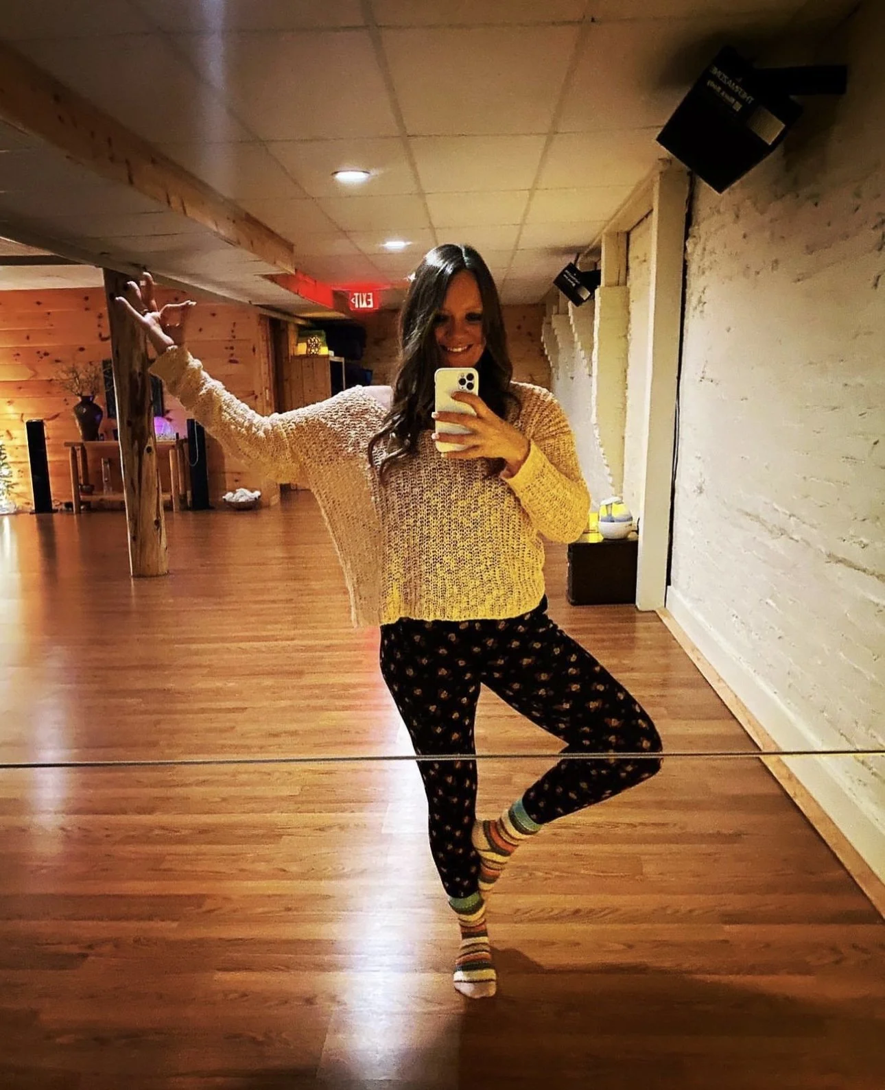 A woman taking a mirror selfie in a wooden-floored room, wearing a yellow knit sweater, patterned pants, and colorful striped socks, with a relaxed pose and a smile.