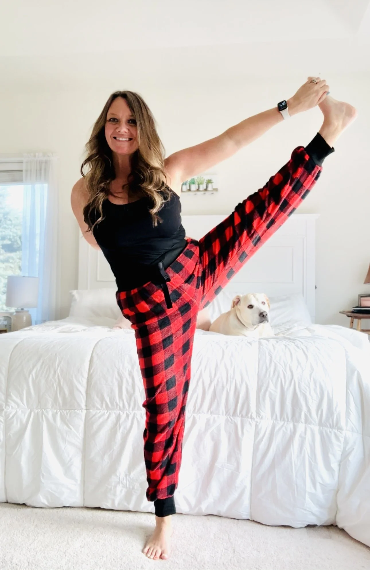 Woman doing a yoga pose on a bed, wearing red and black plaid pajama pants and a black tank top, with a dog sitting on the bed nearby.
