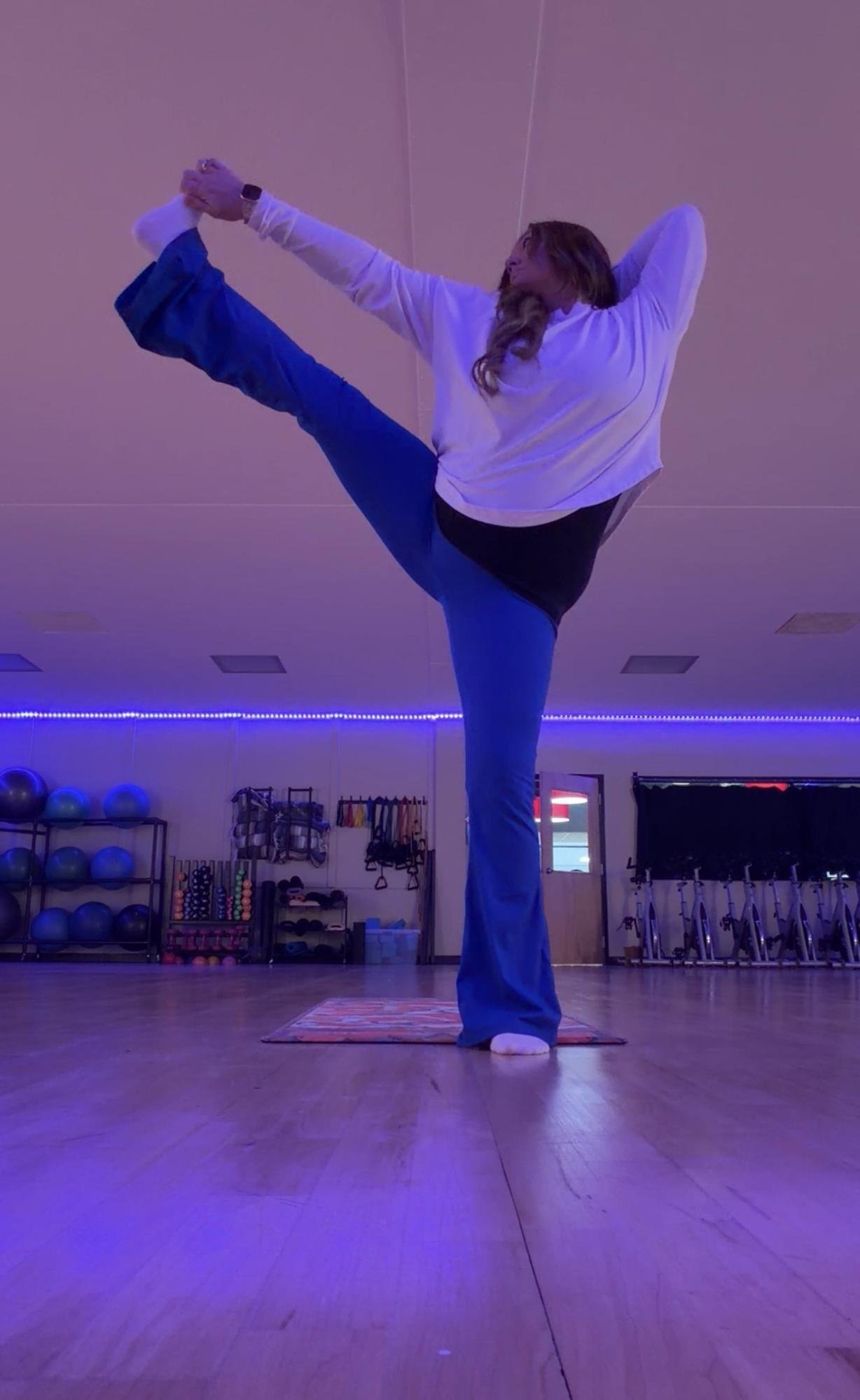 A woman practicing yoga in a gym studio, standing on one leg with her other leg extended upward and held with her hand, while looking to the side and smiling. The studio has exercise equipment and a purple lighting ambiance.