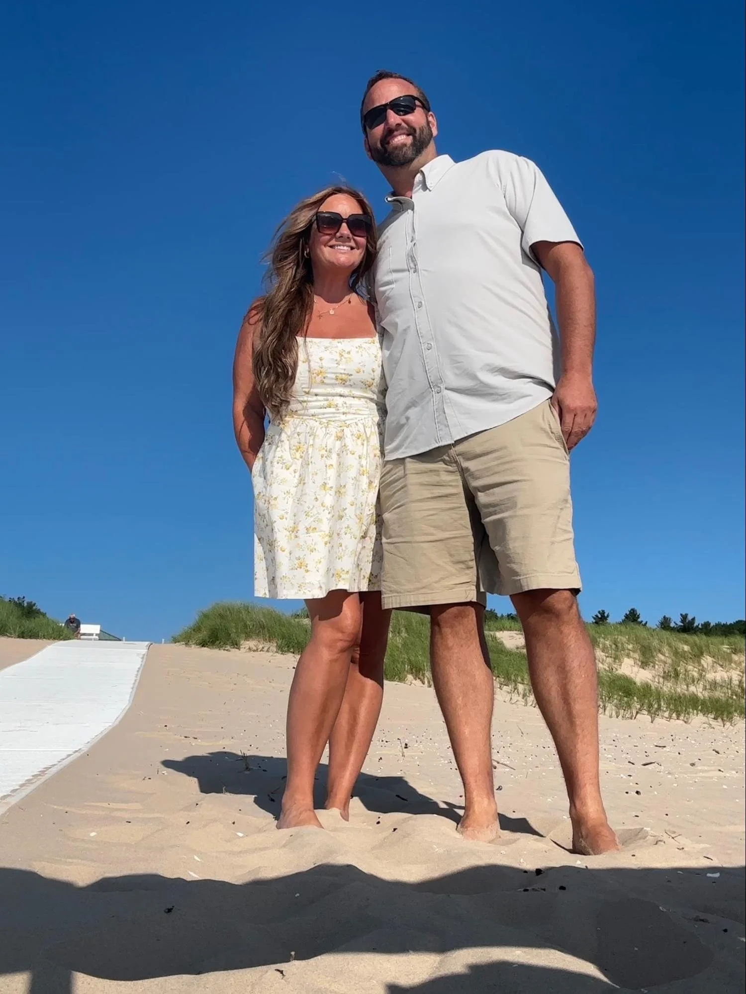 A couple standing barefoot on a sandy beach, smiling at the camera. The woman has long wavy hair and is wearing a white floral sundress and sunglasses. The man has a beard, short hair, and is wearing sunglasses, a light-colored short-sleeved shirt, a