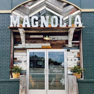 Exterior of Magnolia Boutique Coffee shop with large white letters spelling 'Magnolia' above the entrance, glass double doors with the boutique's name, potted plants on either side, and rustic wooden decor.