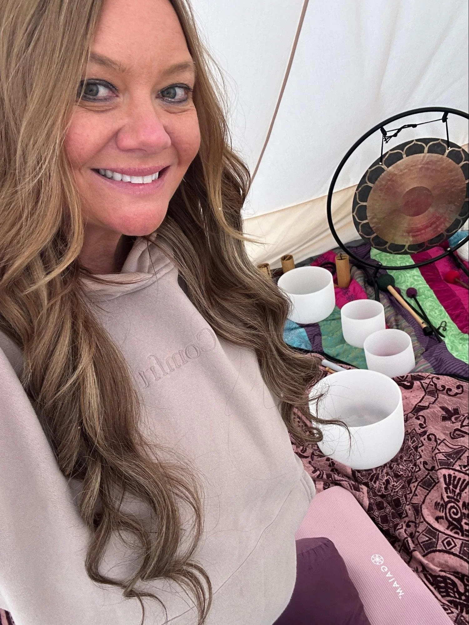 A woman smiling in a tent with singing bowls and a gong on the floor, and a colorful patterned cloth.