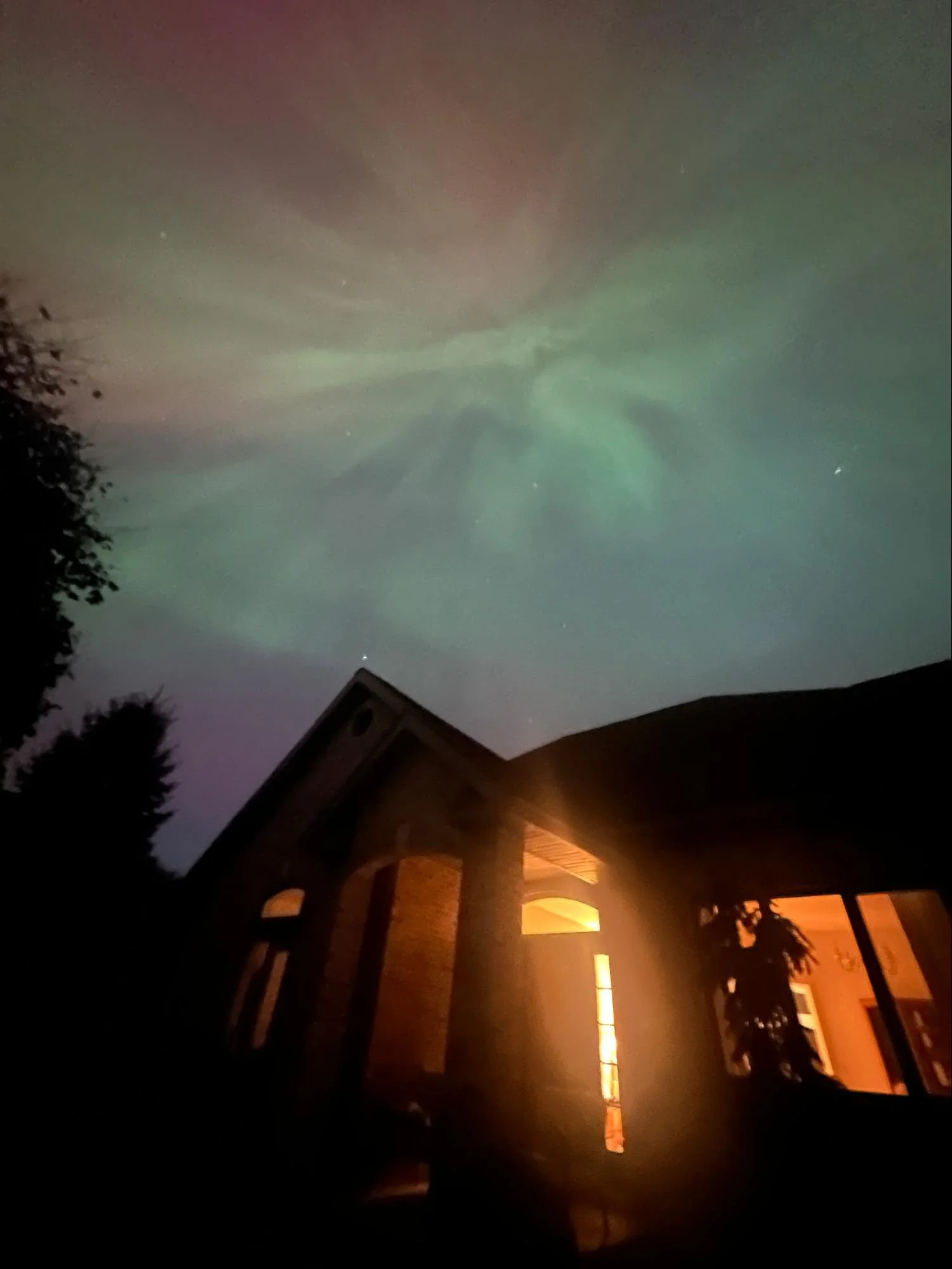 Night sky illuminated by northern lights over a residential house with trees.