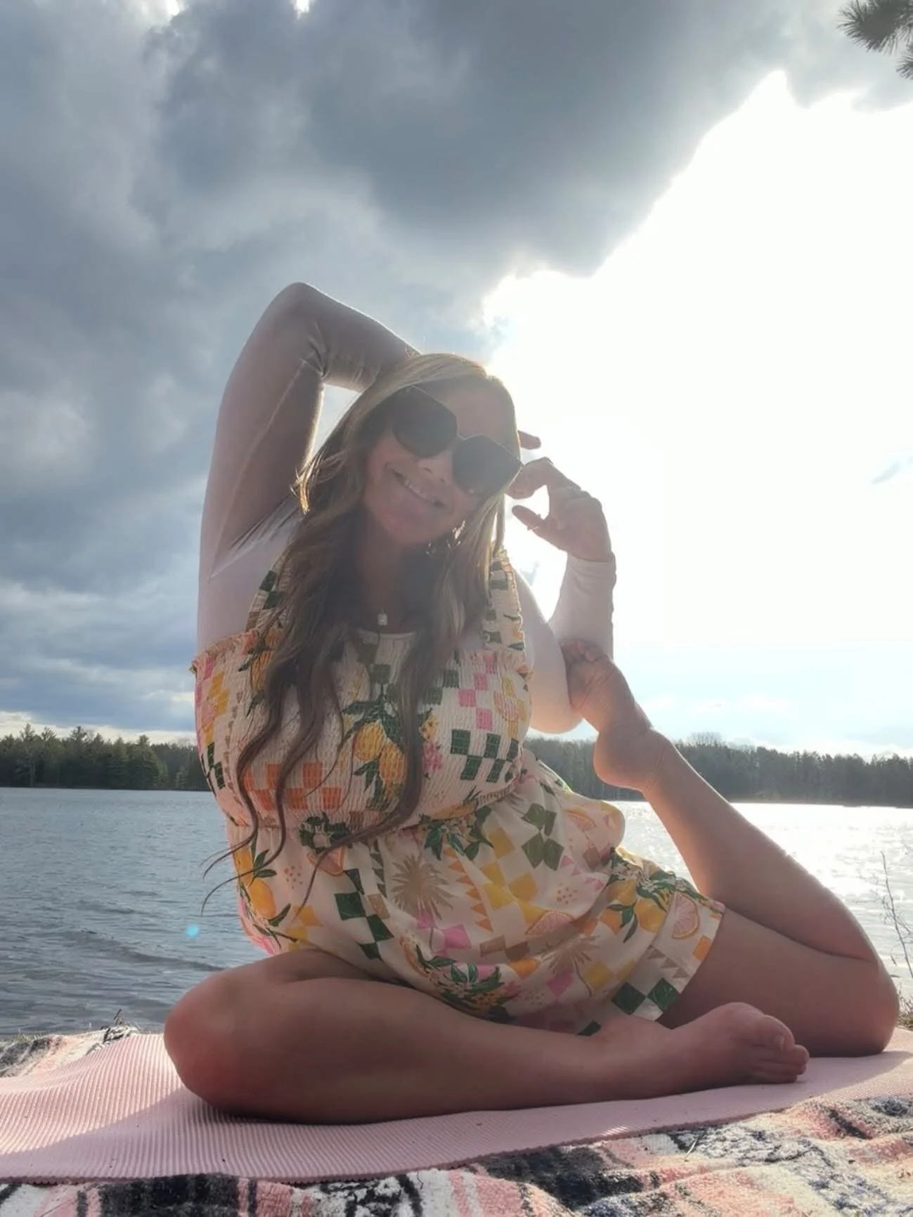 Woman practicing yoga outdoors on a pink mat near a body of water, wearing sunglasses and a floral dress, with dark clouds and the sun in the sky.