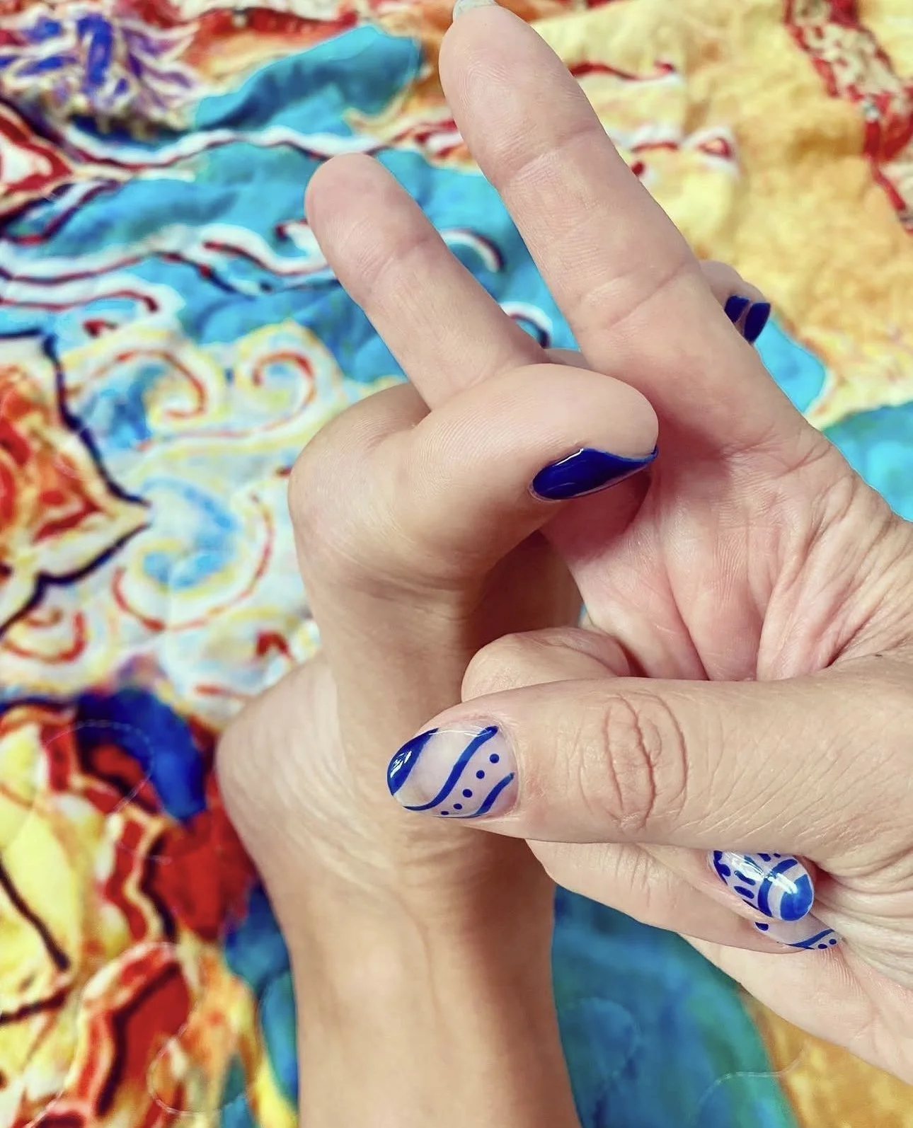 Close-up of a hand with blue and white decorative nail art, held in front of a colorful, abstract-patterned fabric background.