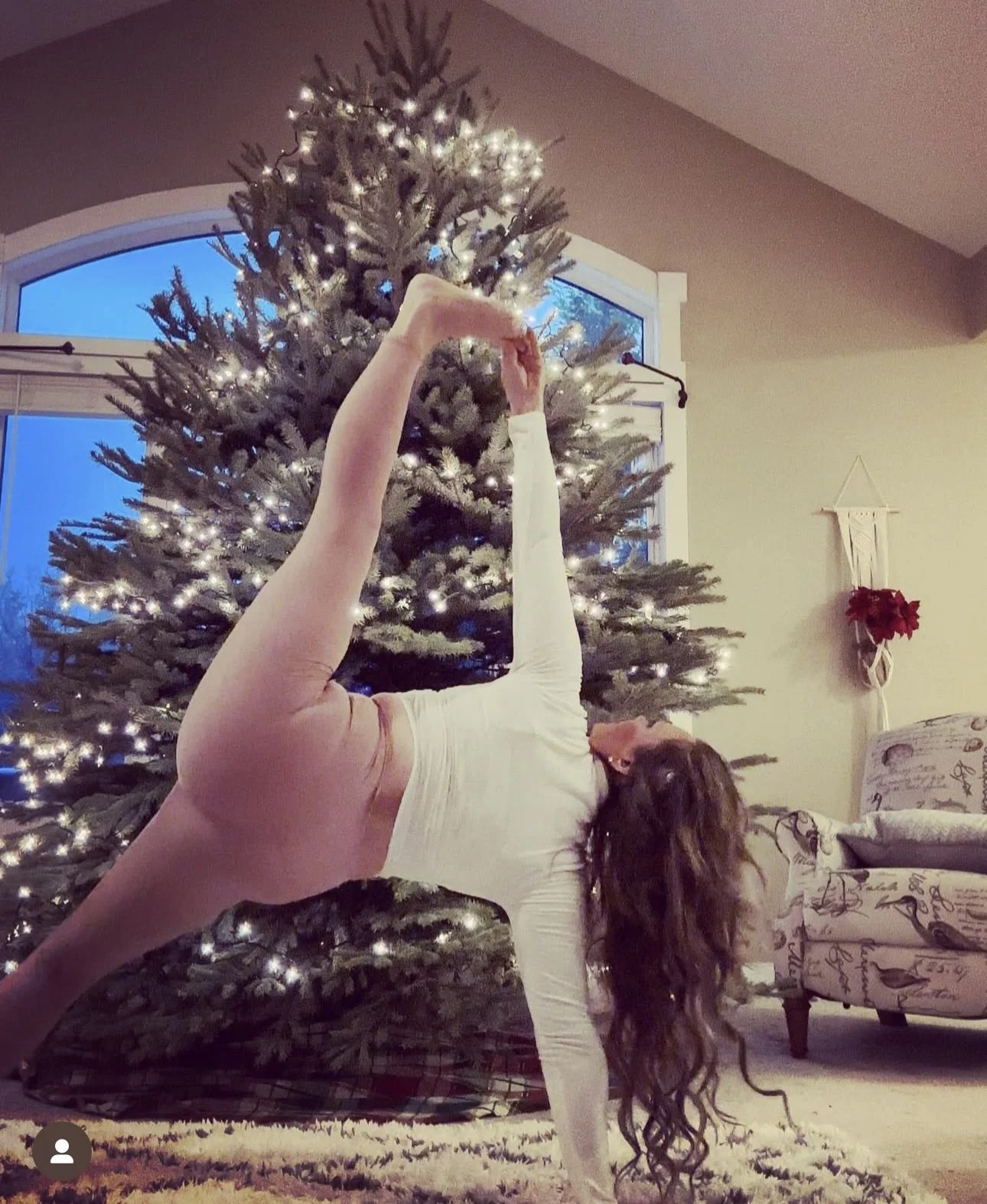 A person doing a yoga pose in front of a decorated Christmas tree with lights, holding their leg with one hand and arching backward, with a dog sitting beside them.