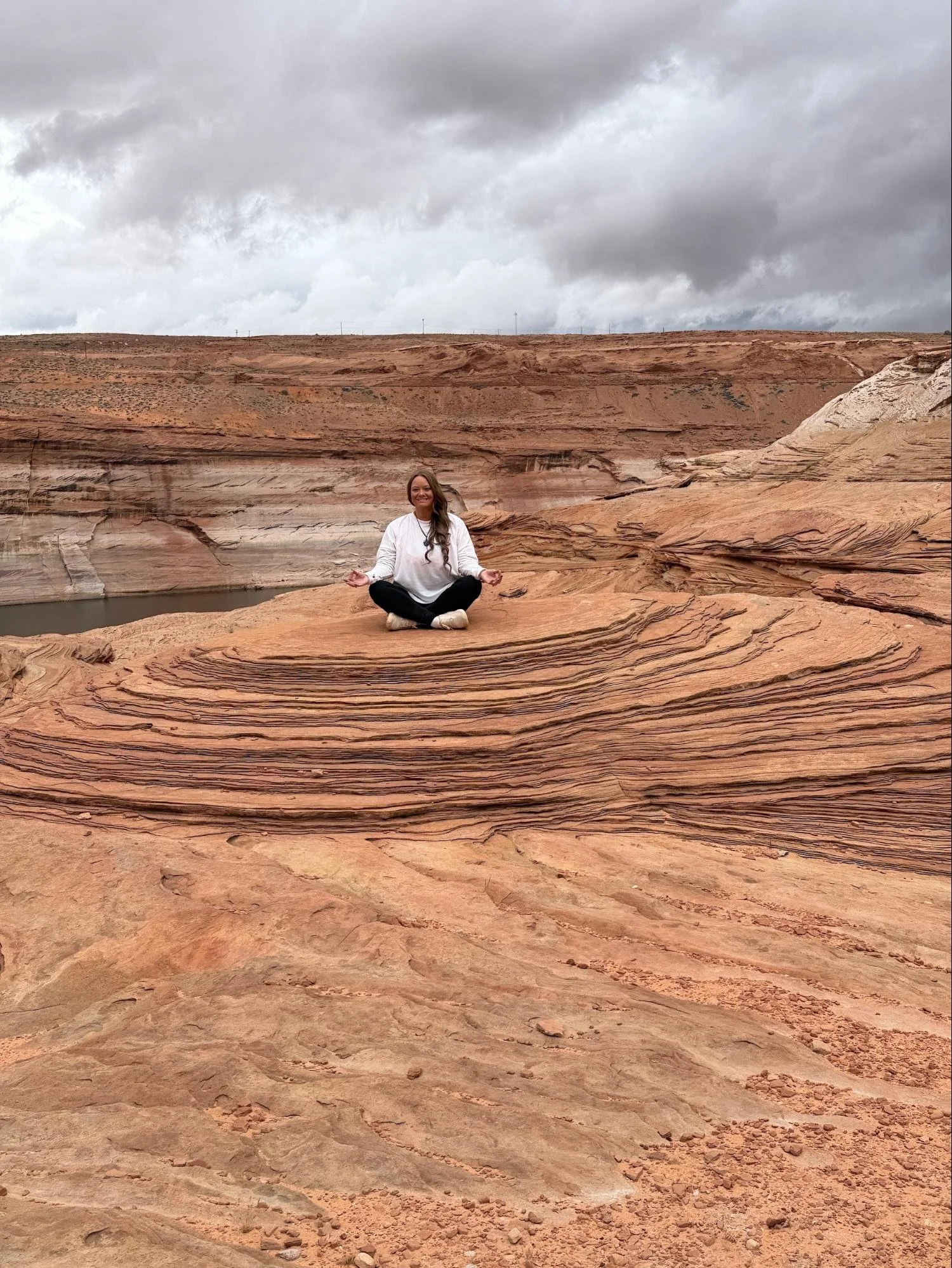A woman meditating in a seated yoga pose on layered red sandstone in a desert landscape under a cloudy sky.
